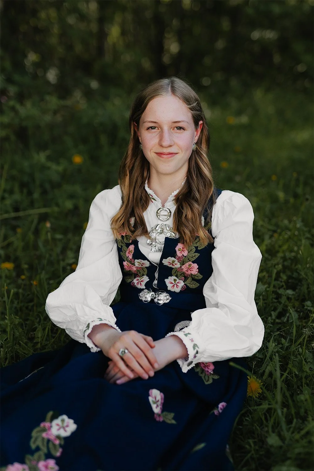 Et portrett av en konfirmant som sitter i gresset utendørs, nær skogen. Hun har på seg Lier-bunad, har hendene i fanget og ser smilende mot kameraet.