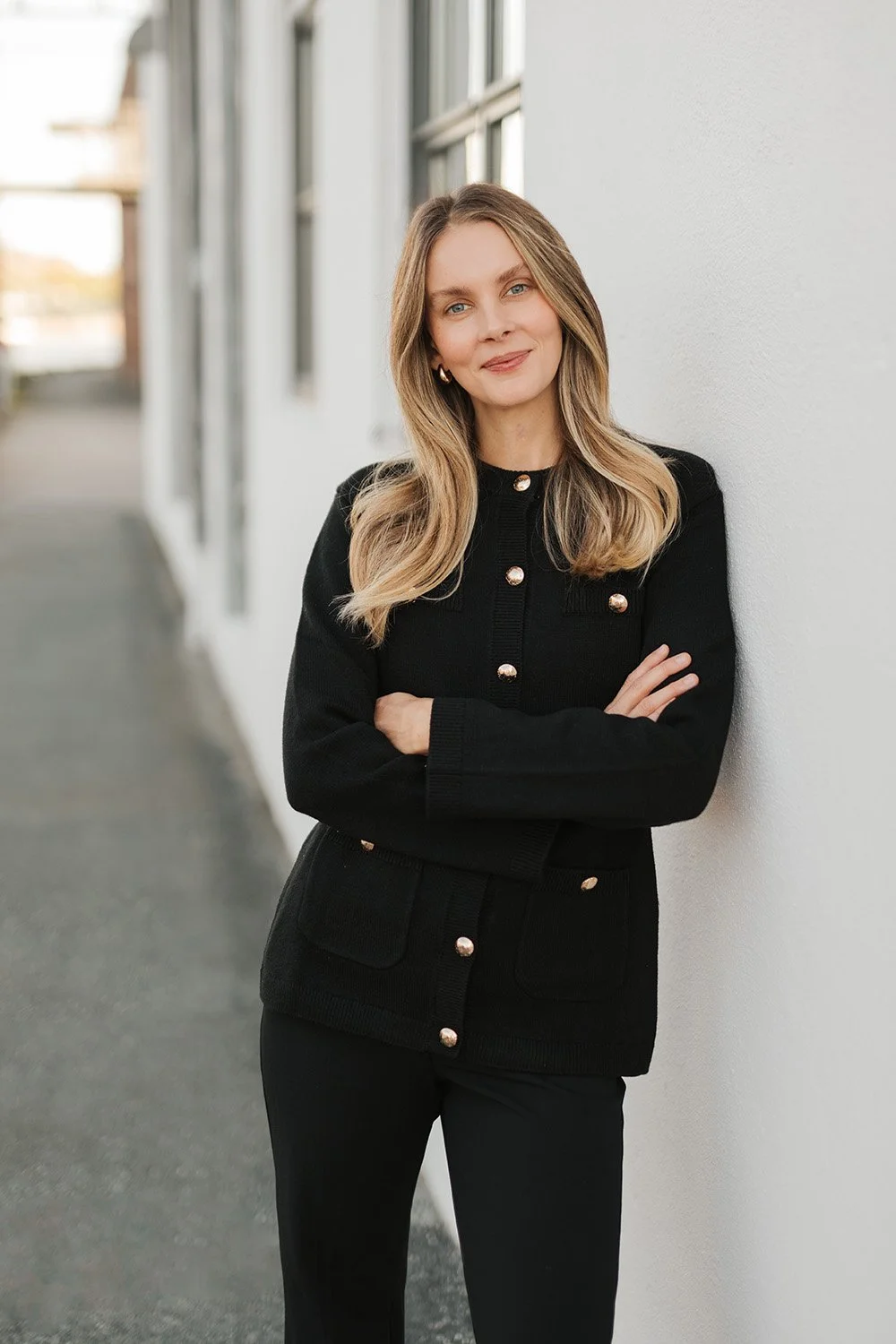 Portrettfotografering: En smilende kvinne med langt bølgete blondt hår og svart jakke med gullknapper, som lener seg mot en hvit vegg.