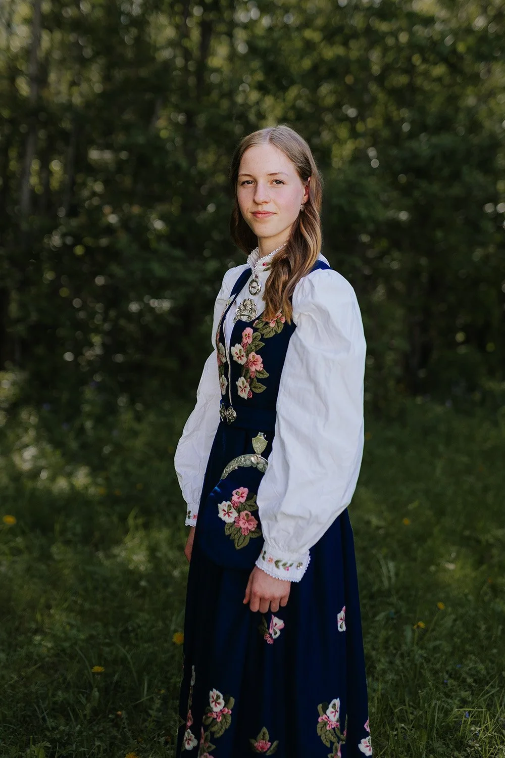 En konfirmant stående i skogkanten, iført en blå Lier-bunad. Hun står vendt litt bort fra kameraet, men ser tilbake over skulderen med et mildt smil.
