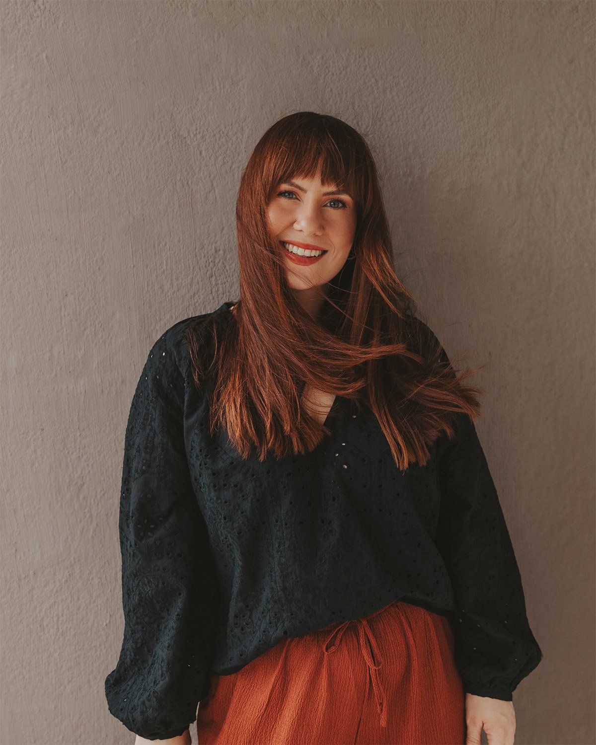 Et smilende portrett av fotograf Tina Bergersen fra Drammen, med rødt hår og pannelugg, foran en grå vegg. Hun har på seg en svart bluse og rødbrune bukser.