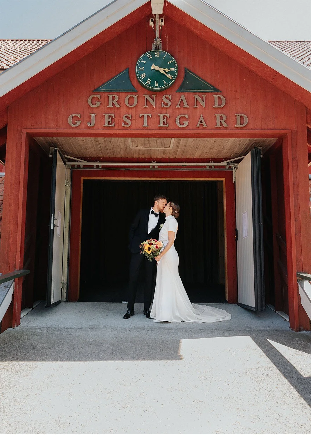 Ett brudepar som kysser foran den røde hovedinngangen på Grøsand Gjestegård i Sætre. Tatt av Tina Bergersen, bryllupsfotograf i Drammen.