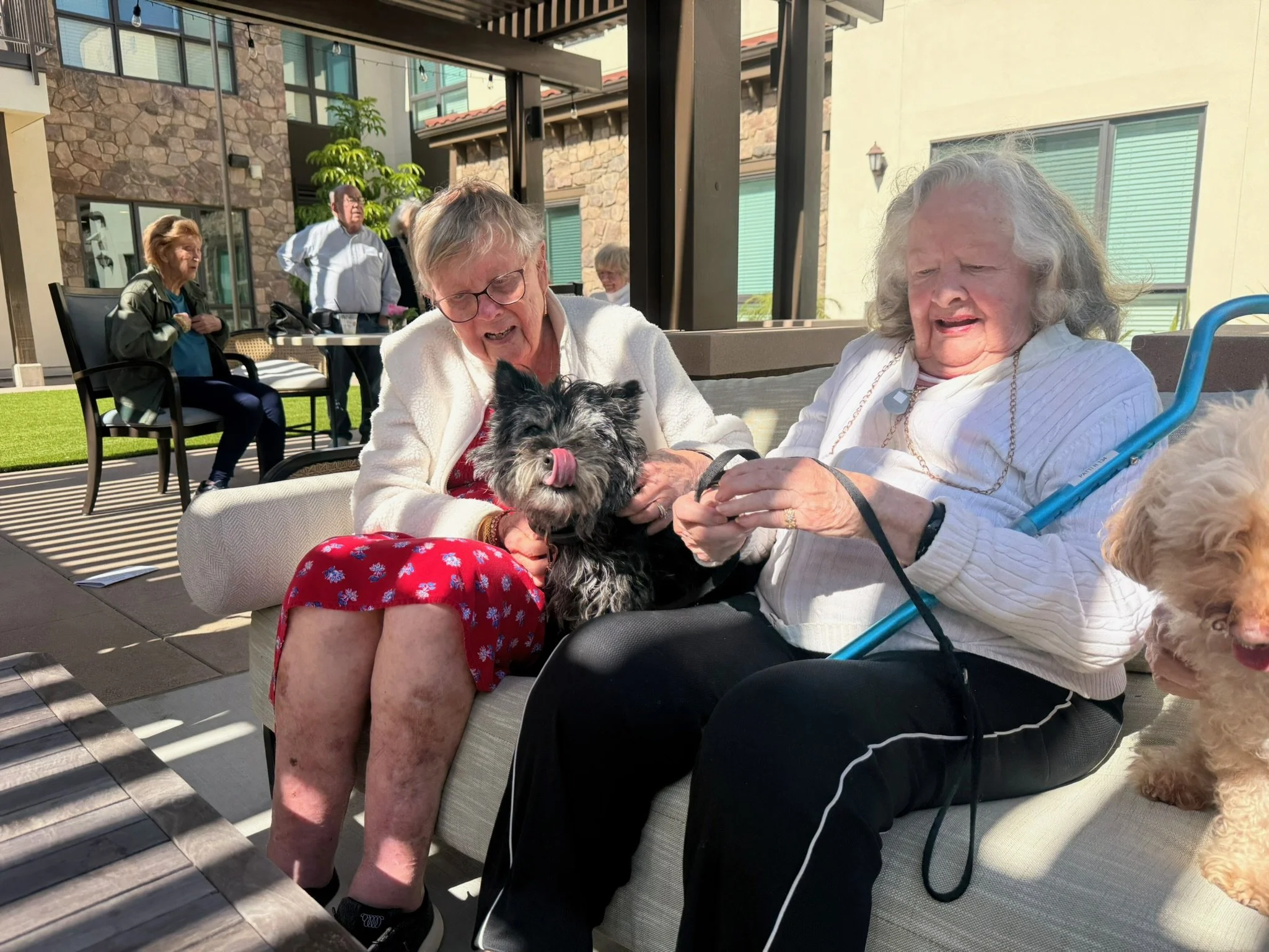 Two senior living residents petting a small dog while seated together in an assisted living community in San Diego