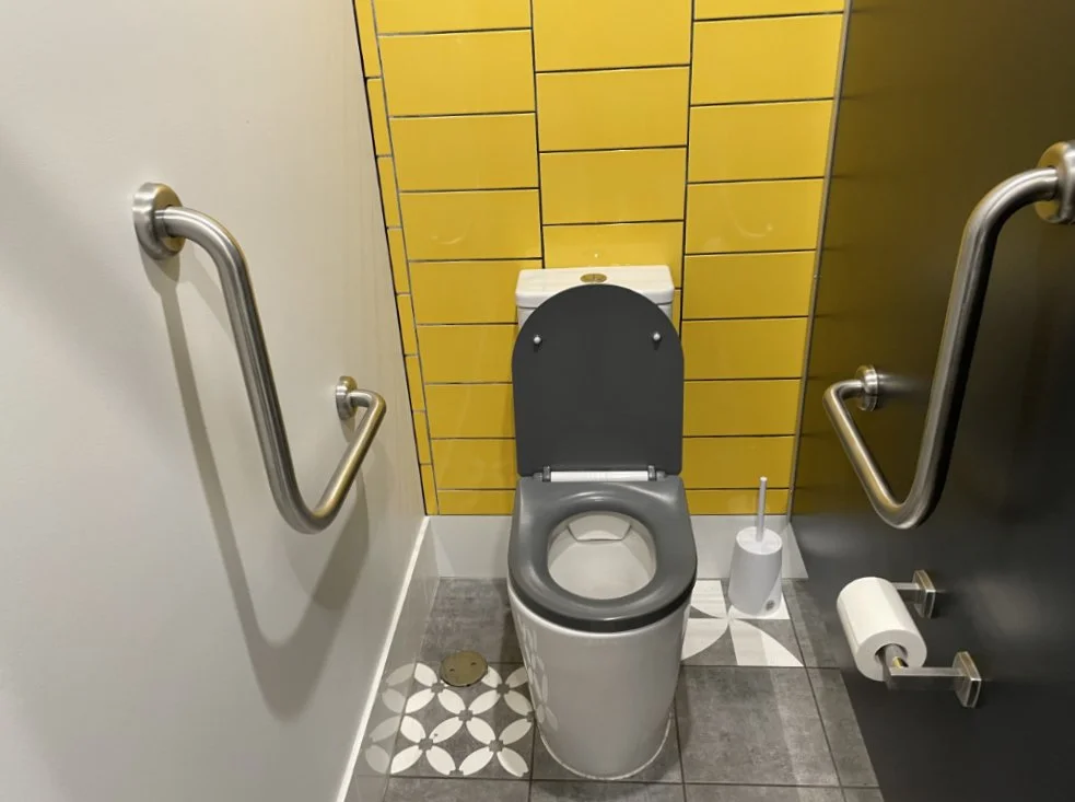 An ambulant toilet with grabrails against a bright yellow tiled wall
