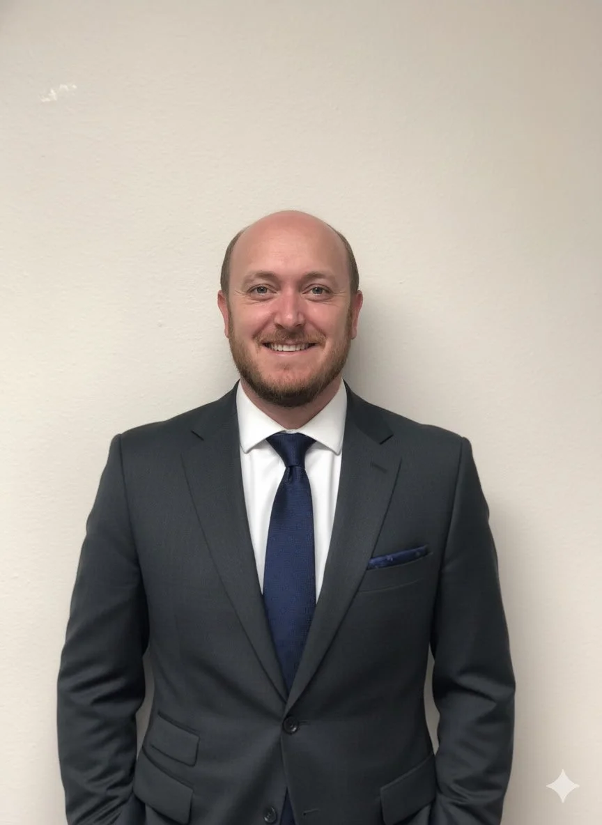 A man in a dark suit, white shirt, and blue tie stands smiling against a plain, off-white wall.