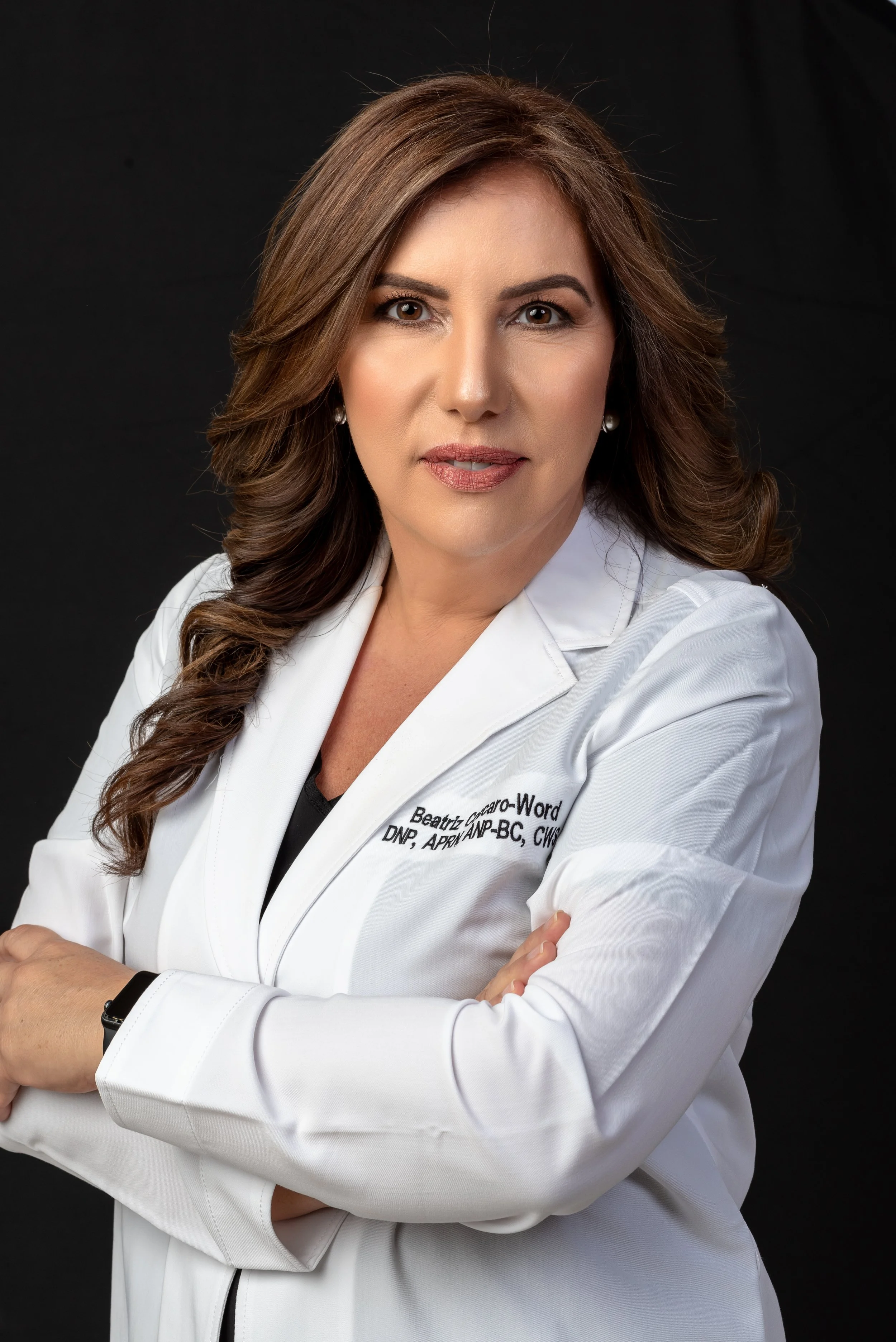 A woman with long brown hair, wearing a white medical coat with arms crossed, against a black background.