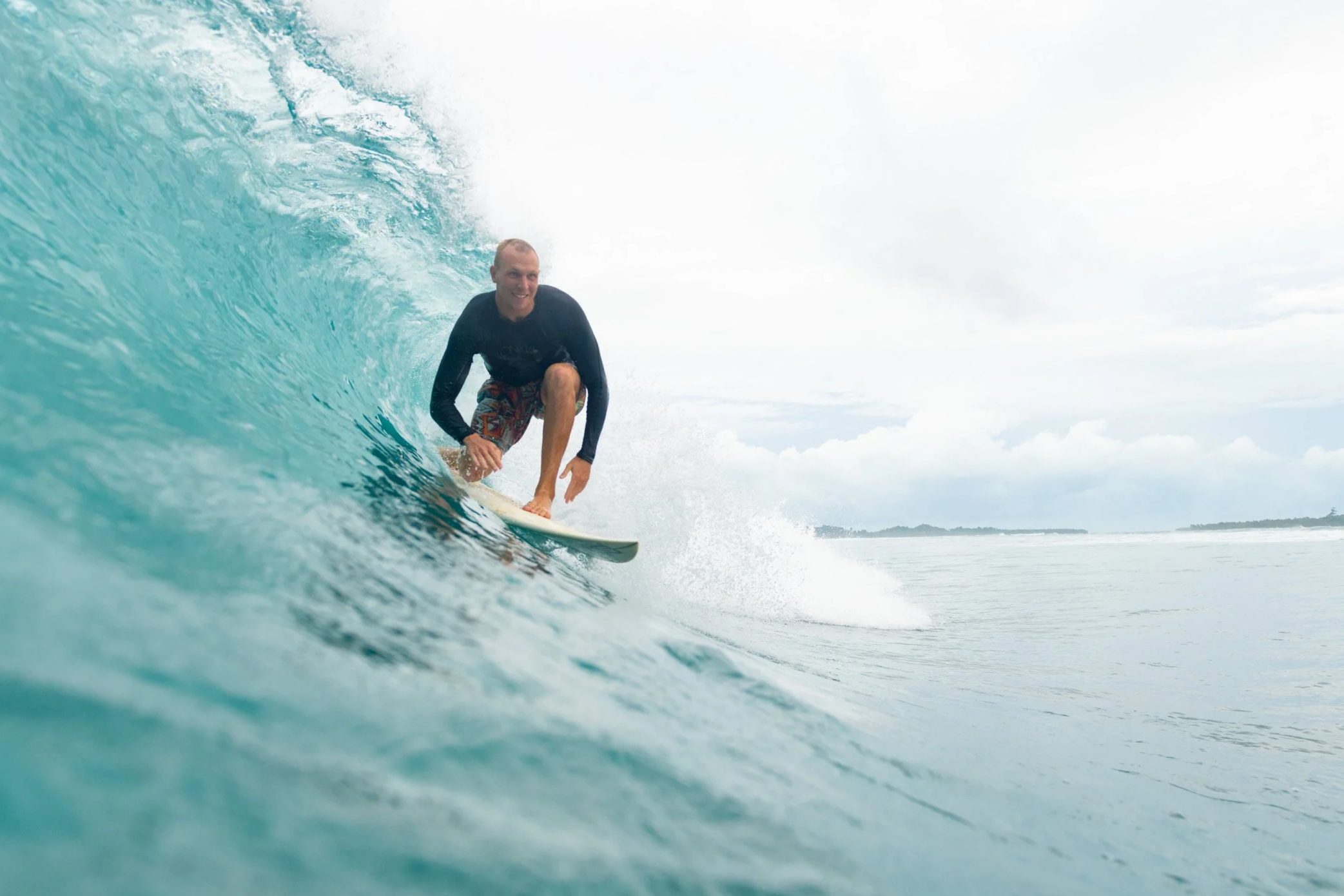 A man surfing on a turquoise wave with a smile, wearing a black wetsuit and patterned swim shorts, during daytime with a cloudy sky.