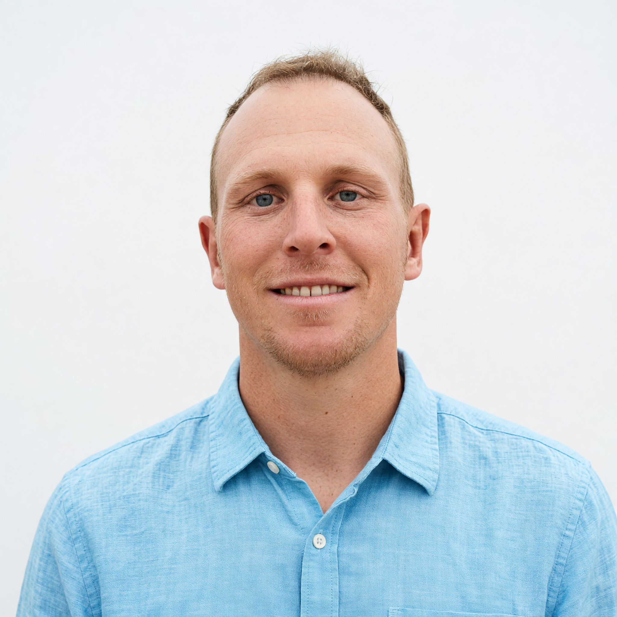 Gold Coast architect - Close-up of a cheerful young man with light skin, blue eyes, and short red hair, wearing a light blue button-up shirt, smiling outdoors against a white background.