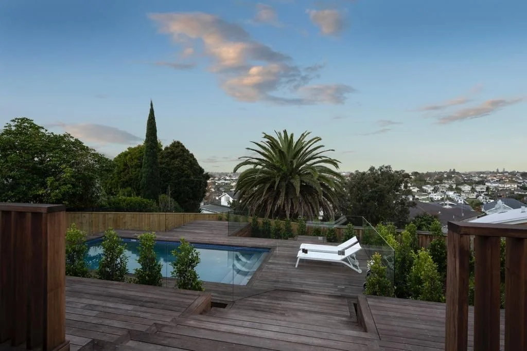 A rooftop deck with wooden flooring, a small swimming pool, a white lounge chair, green bushes, and a large palm tree, overlooking a neighborhood with houses and trees under a partly cloudy sky.