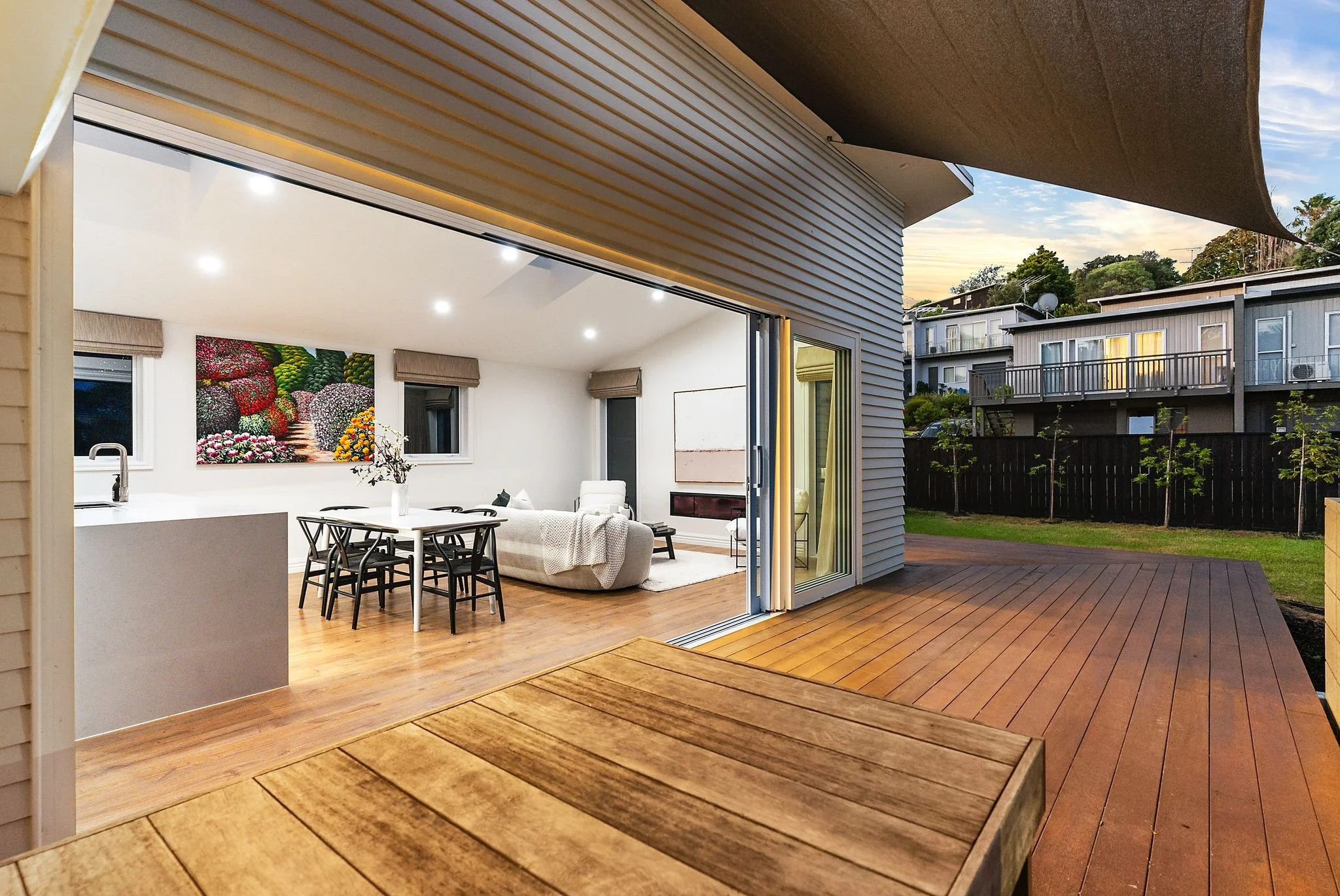 View of a modern living room inside a house with sliding glass doors opening to a wooden deck and backyard, evening sky in background.