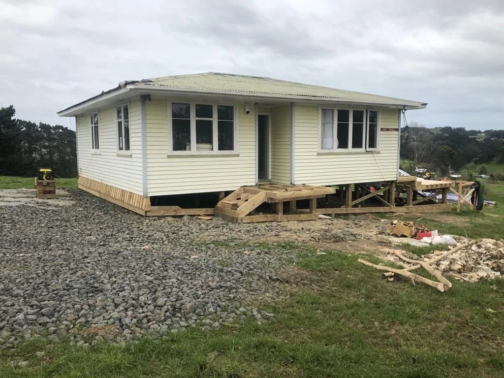A house under construction with a raised foundation, yellow siding, and wooden stairs near the entrance, surrounded by construction materials and gravel.