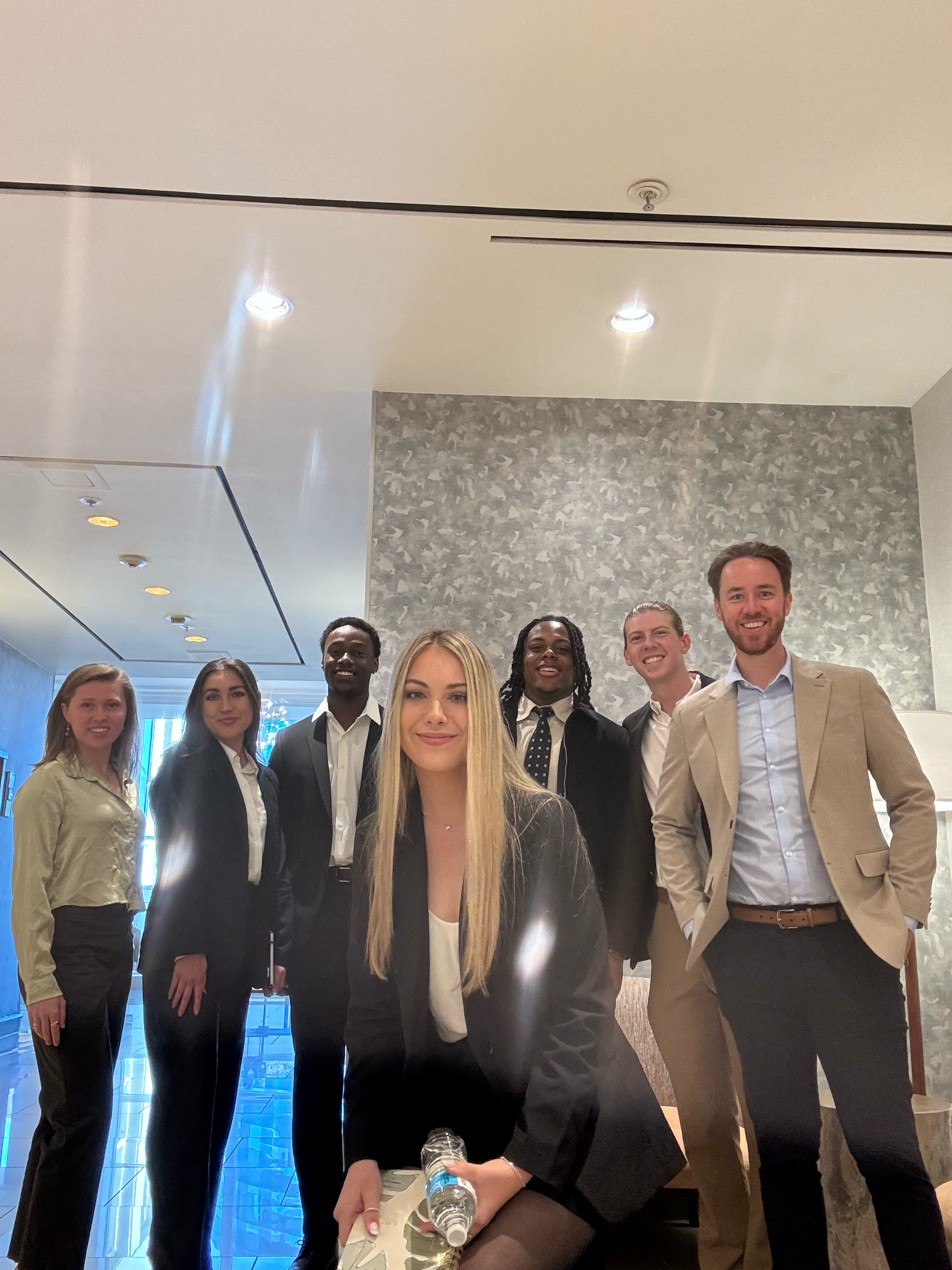 Group of seven young professionals in business attire, standing inside a modern building with glass windows, smiling at the camera.