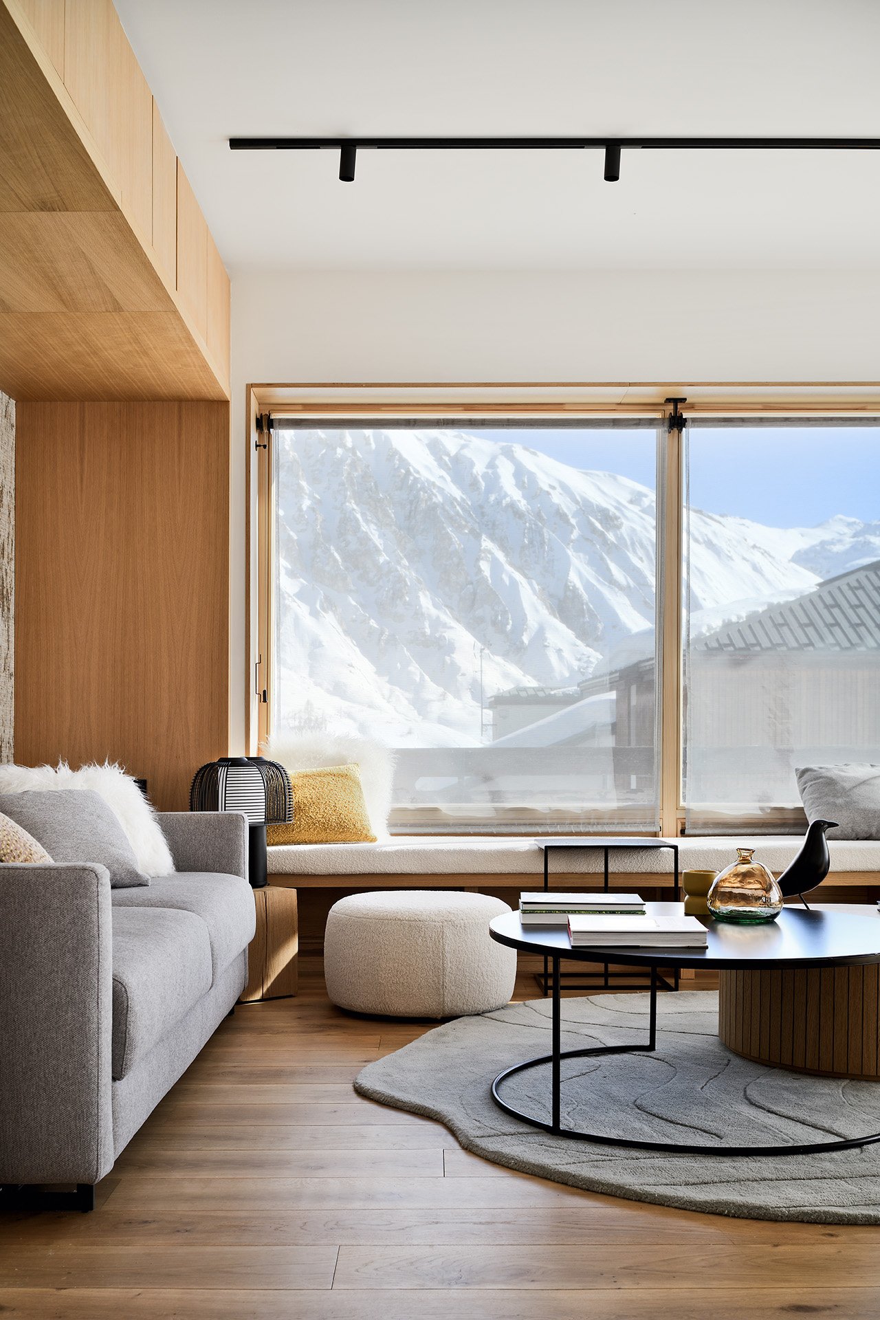 Salon moderne avec vue sur une montagne enneigée, mobilier en bois clair, canapé gris clair avec coussins blancs et colorés, table basse ronde noire, tapis beige, décor minimaliste avec objets en verre et en céramique.
