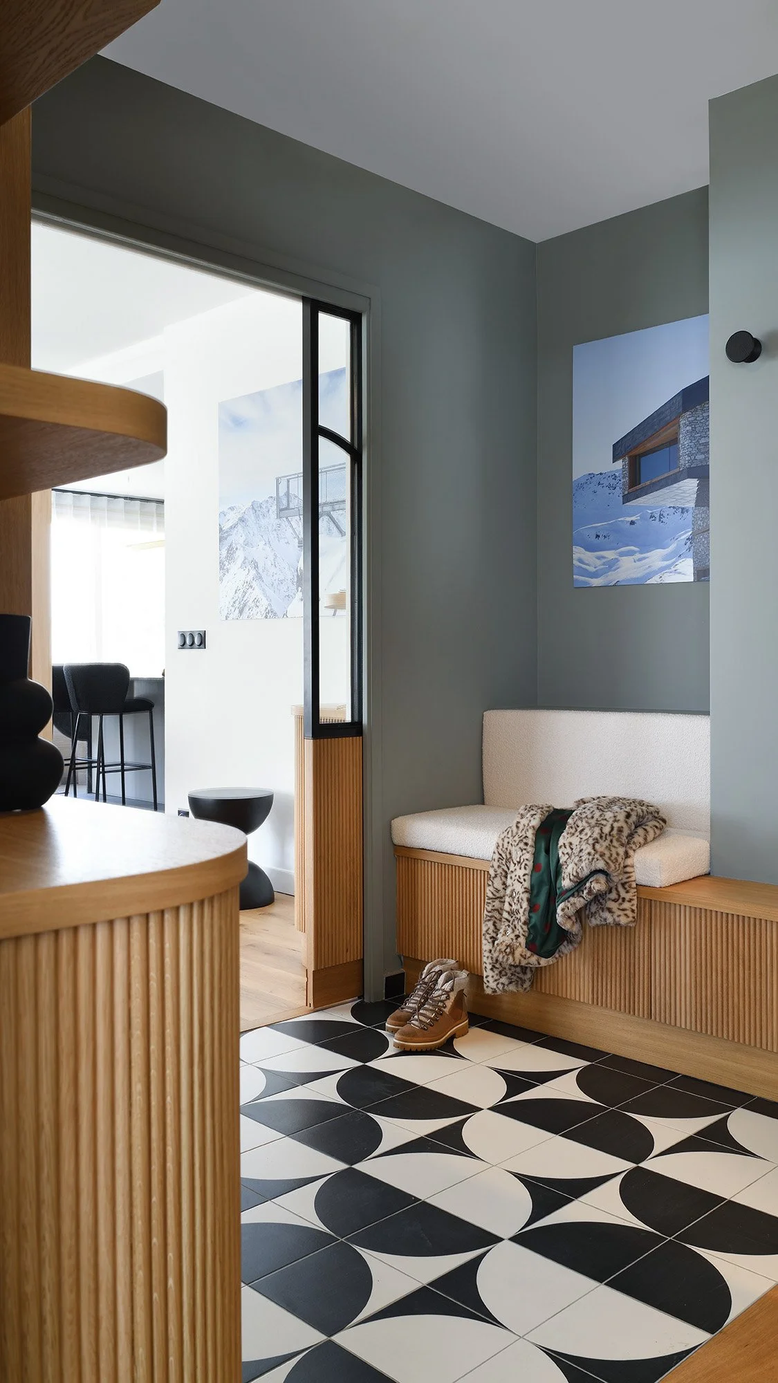 Un coin d'entrée avec un banc en bois, un tapis noir et blanc à motifs géométriques, des bottes, un pull et une écharpe posés sur le banc, et un tableau représentant une maison dans un paysage enneigé sur le mur.