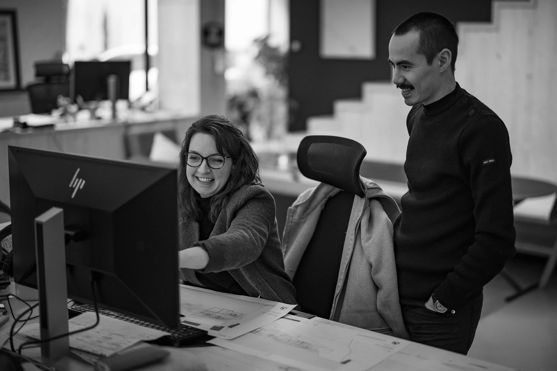 Deux personnes dans un bureau, une femme assise devant un ordinateur souriant, un homme debout à côté regardant l'écran, tous deux en train de discuter et de sourire.