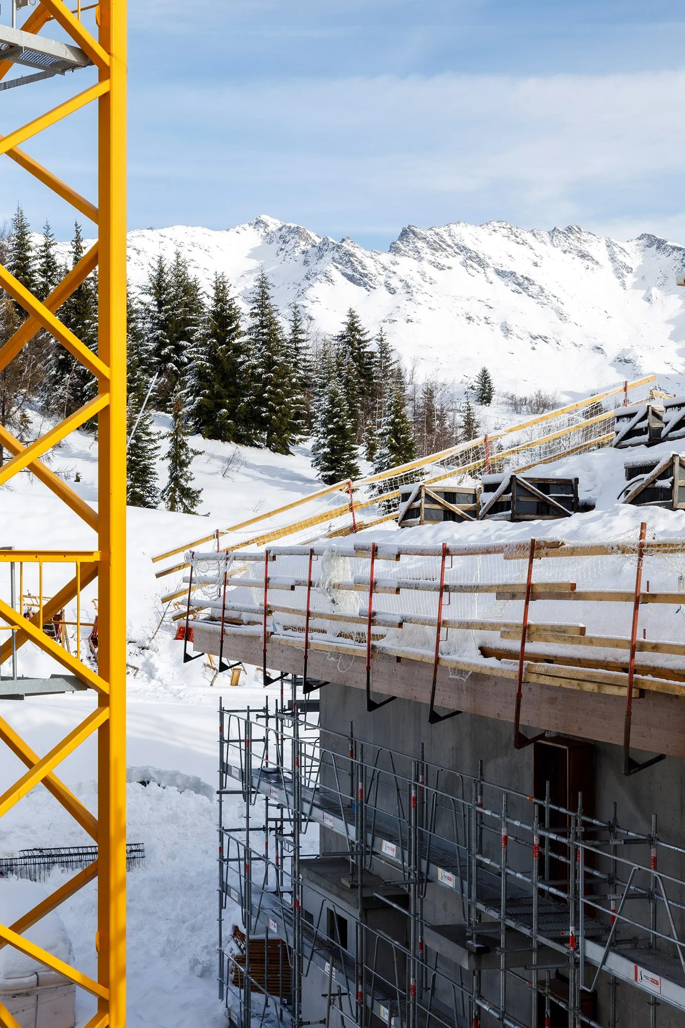 Construction d'un bâtiment en zone enneigée avec échafaudages, au milieu de montagnes enneigées et d'arbres.