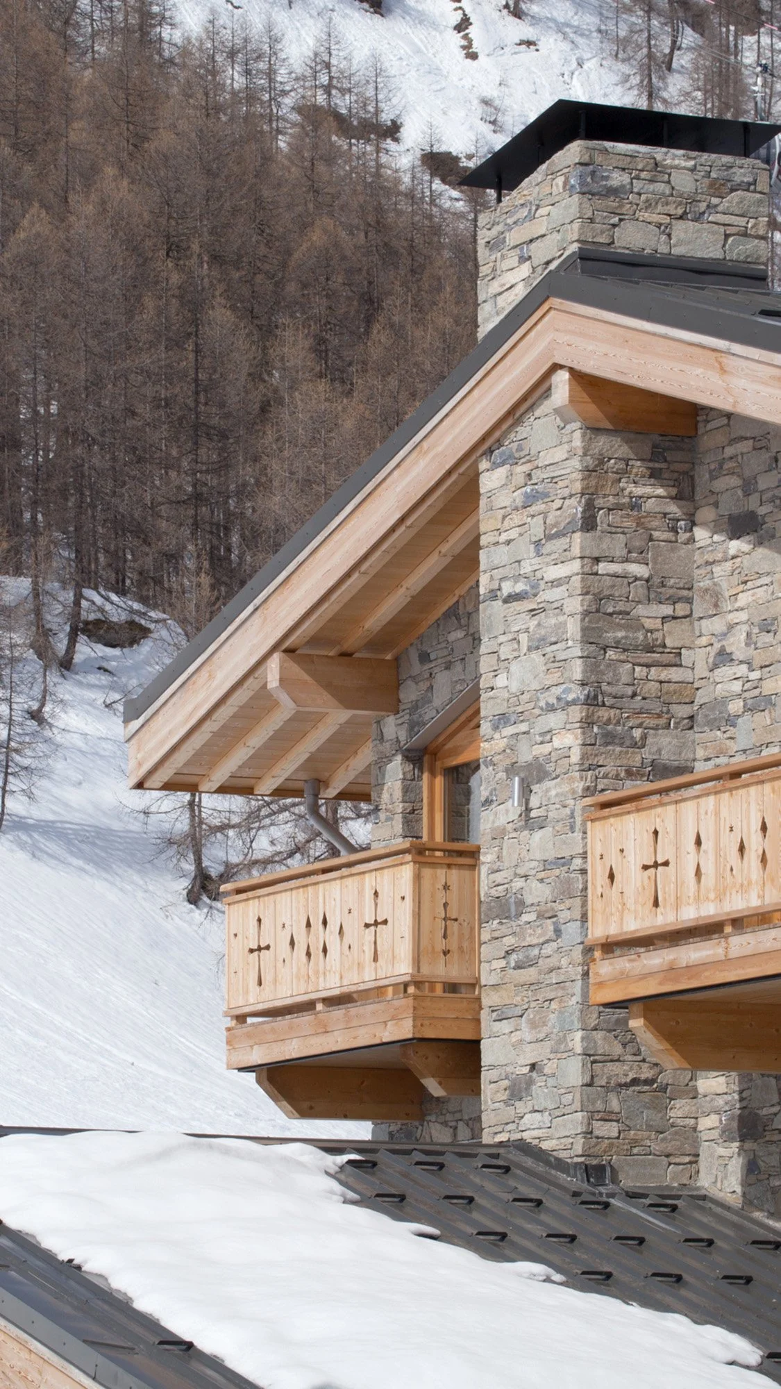 Façade d'une maison en pierre avec balcons en bois, dans un paysage enneigé, avec des arbres et une montagne en arrière-plan.