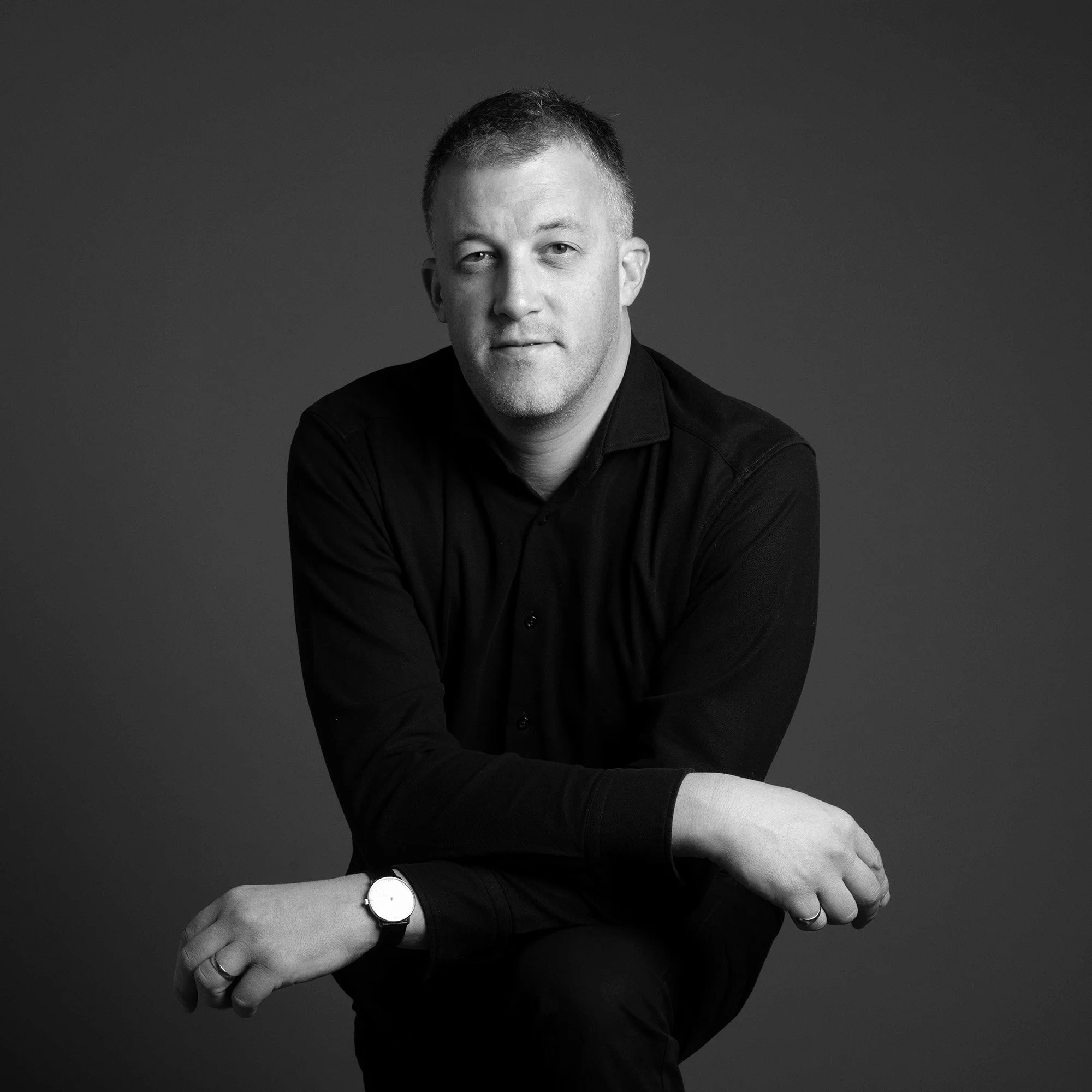 Portrait en noir et blanc d'un homme assis, portant une chemise noire, regardant vers l'appareil, avec un fond gris.