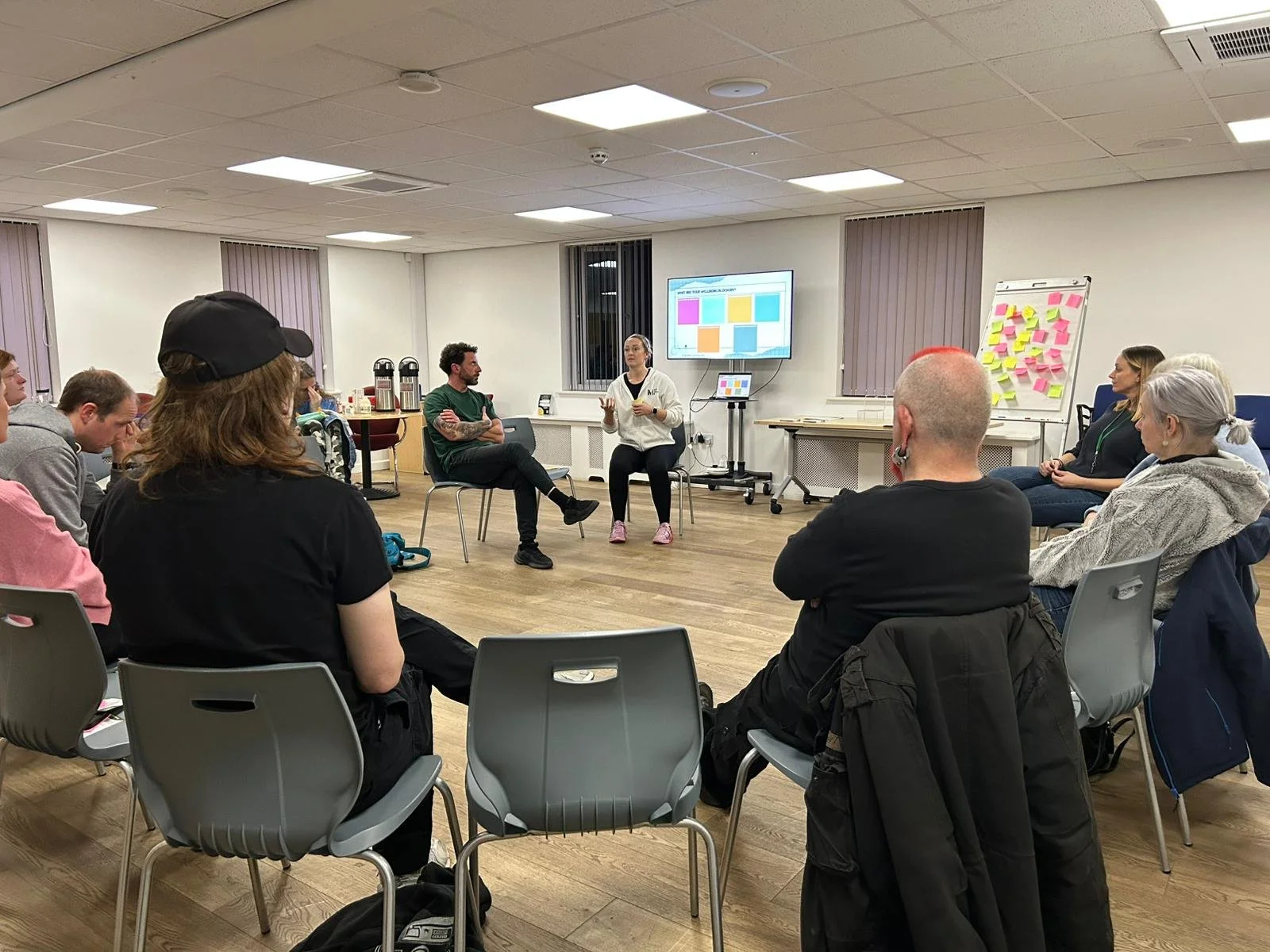 A group of people sitting in a circle in a conference or workshop room, listening to a woman speaking in front of a screen displaying colorful blocks and a whiteboard with pink sticky notes.