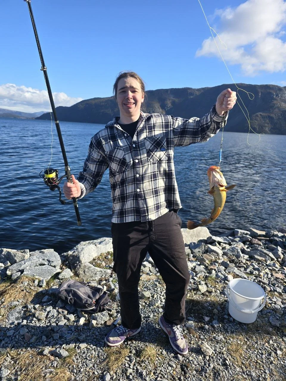 A young person standing on rocks by a lake, holding a fishing rod in one hand and a fish in the other, with mountains and blue sky in the background.