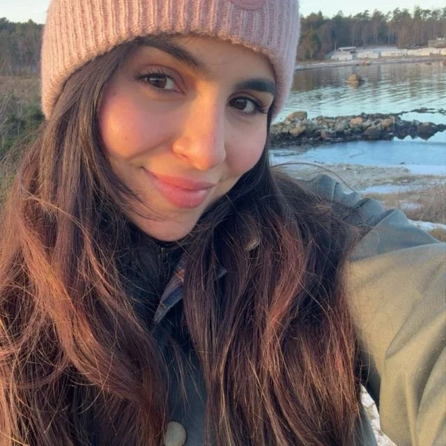 Young woman with long brown hair smiling outdoors near water, wearing a pink knit beanie and a jacket. An intern at the ICT4COP Center researching community-based policing and human security solutions.