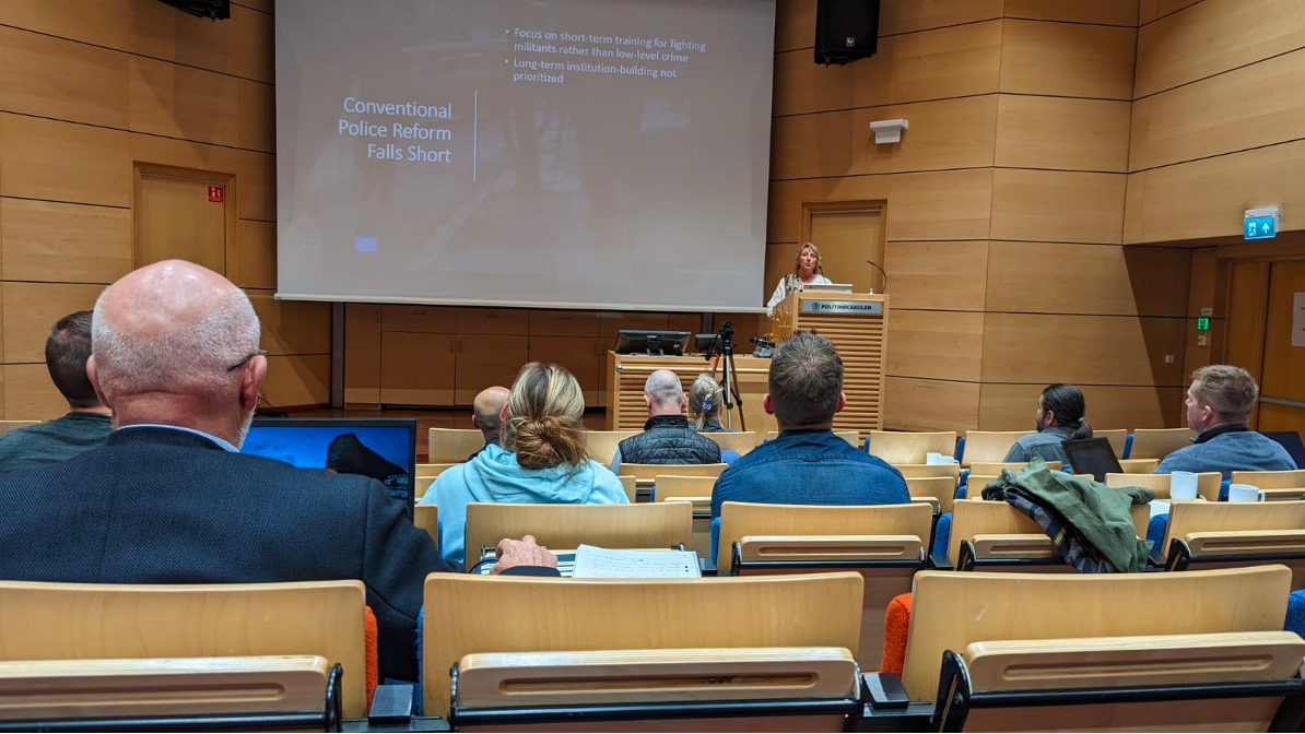 Ingrid Nyborg giving a presentation at a conference or lecture hall with a slide titled "Conventional Police Reform Falls Short." The audience is seated, facing the stage with some people taking notes or using laptops.
