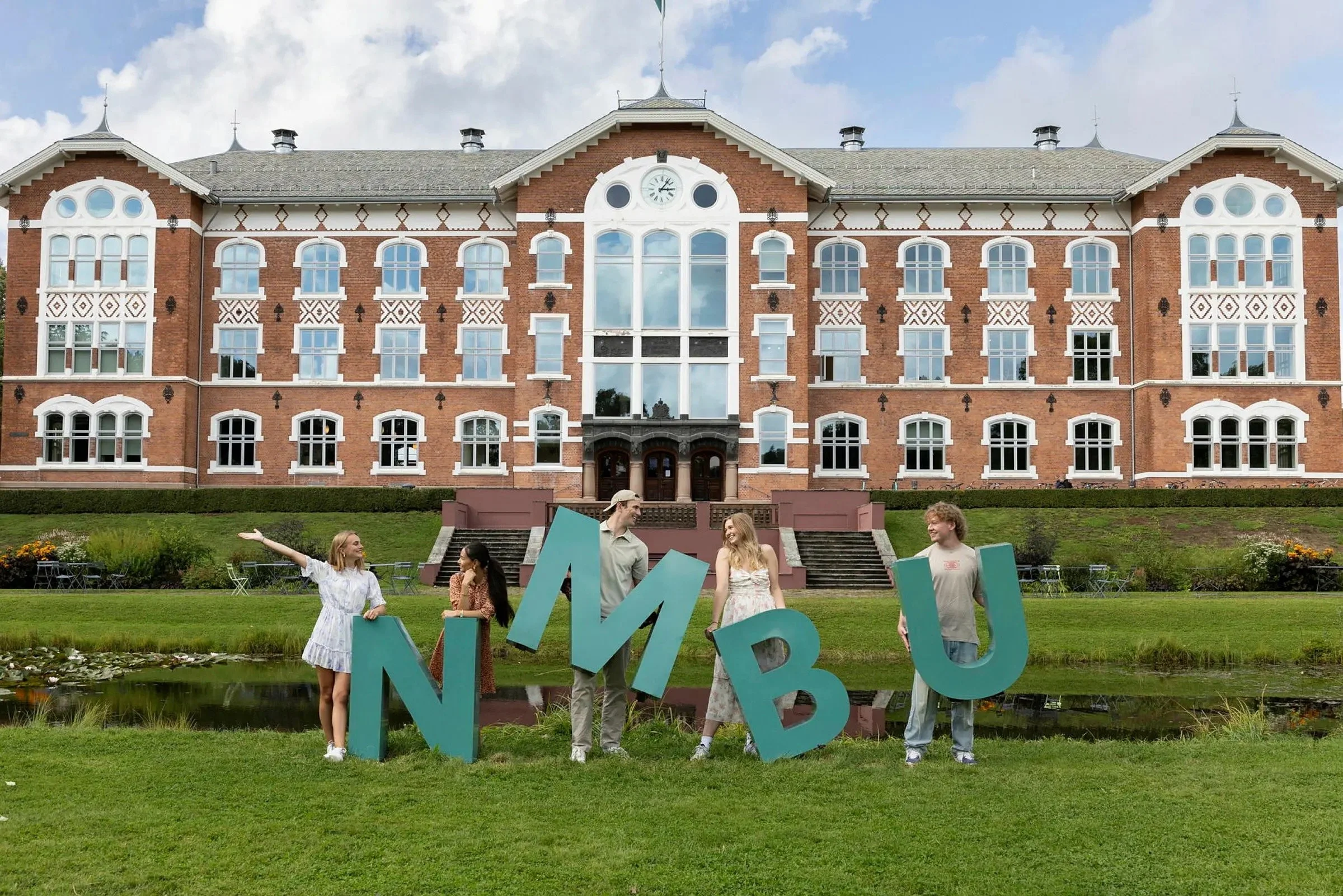 A group of people standing on a lawn in front of a large, historic brick building with many windows and a clock at the top. Some of the people are holding large blue letters, and they are smiling and talking. The scene is outdoors during the day with a partly cloudy sky.