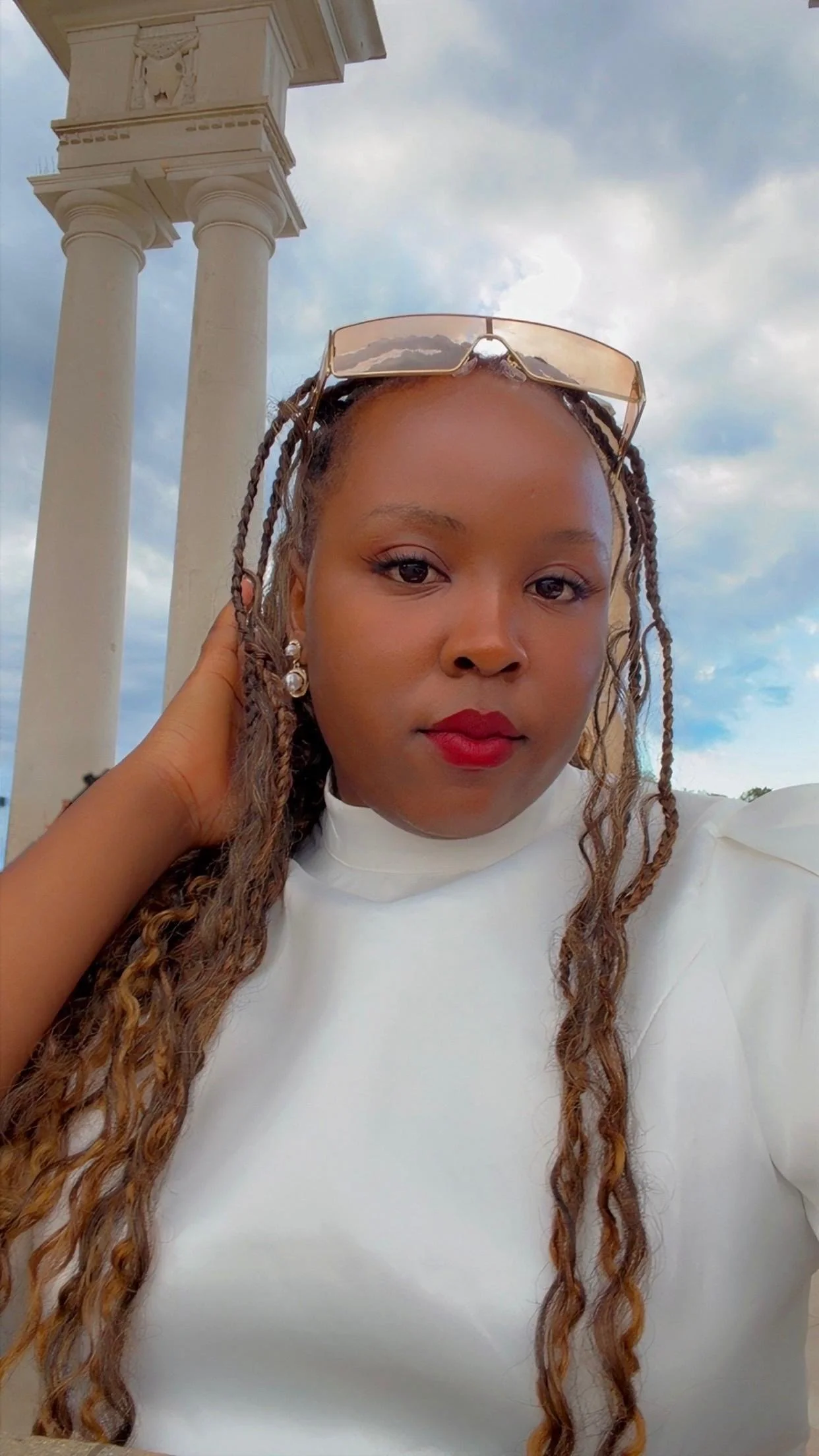 Close-up of a woman with braided hair wearing sunglasses on her head, red lipstick, pearl earrings, and a white top, with a background of cloudy sky. An intern at the ICT4COP Center researching community-based policing and human security solutions.