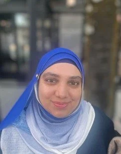 A woman wearing a blue hijab, smiling outdoors with a blurred urban background. An intern at the ICT4COP Center researching community-based policing and human security solutions.