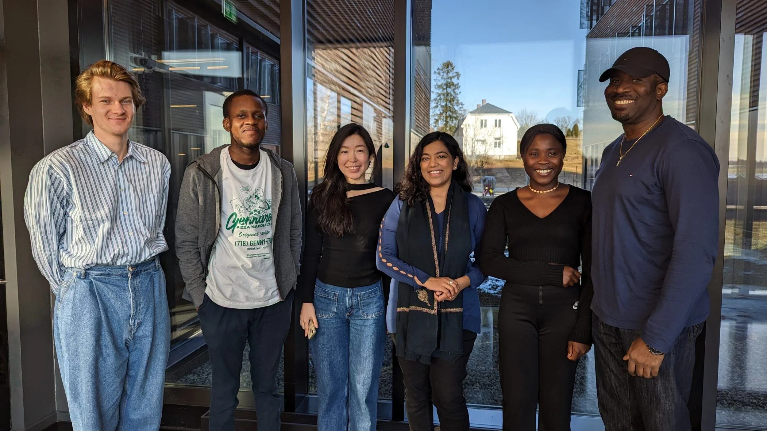 A group of six diverse interns standing together indoors in front of a glass window, smiling at the camera.