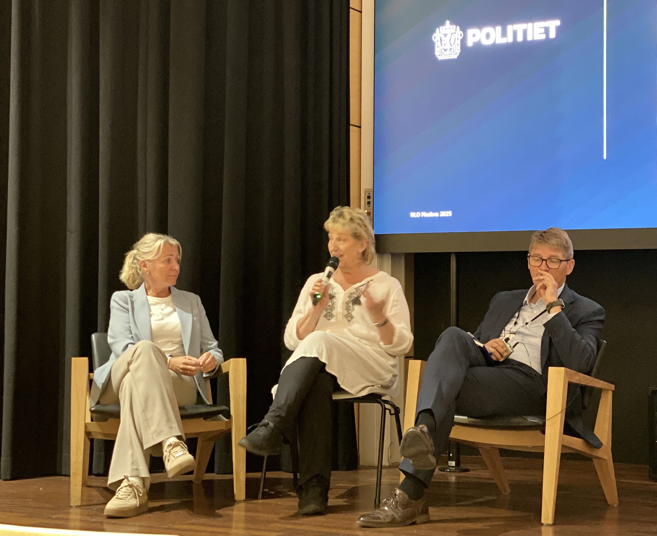 Four people seated on stage during a panel discussion or interview, two women and two men, with a large screen behind them displaying the word 'POLITIET' and a crown logo, indicating it is an event related to the police in Norway.