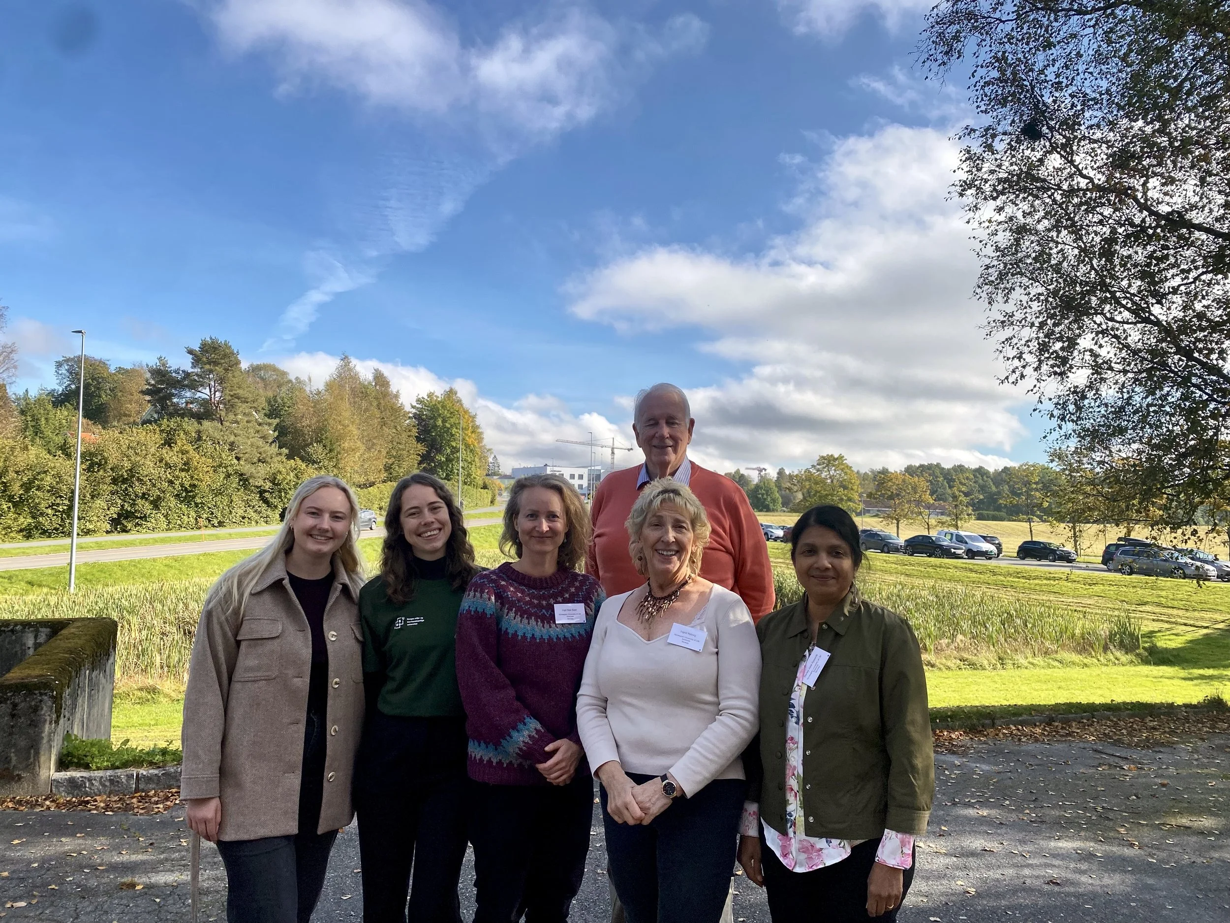 Group of seven diverse researchers standing outdoors in a park with trees and a parking lot in the background, smiling for the camera under a blue sky with scattered clouds.