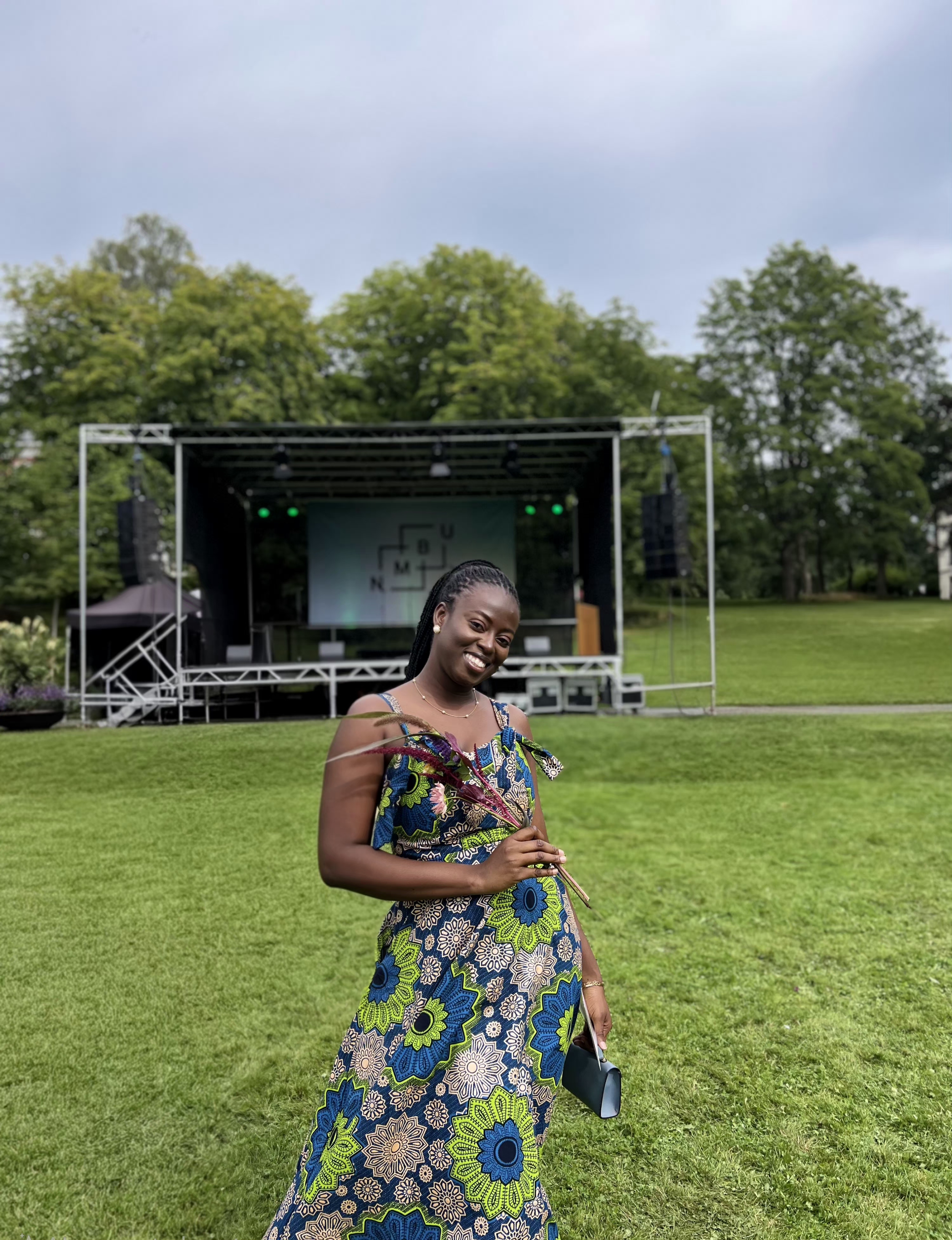 A woman with braided hair wearing a colorful, patterned dress holding a small black purse and smiling, standing on a grassy lawn with a stage, large trees, and a cloudy sky in the background.
