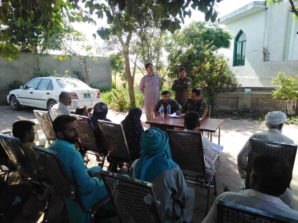 A group of people attending a meeting outdoors with a police officer, some seated and some standing, in a shaded area with trees and a building in the background.