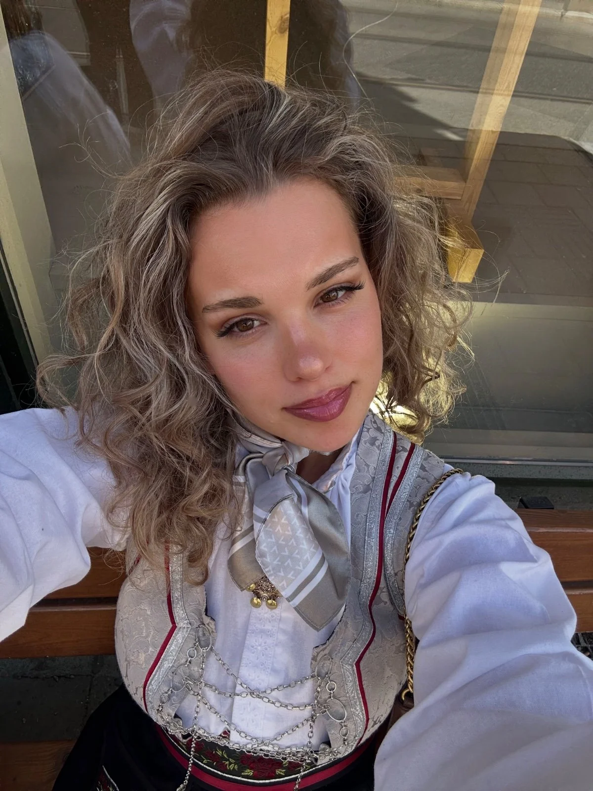 A young woman with curly light brown hair and makeup, wearing a traditional embroidered white blouse with a grey satin neck scarf and silver jewelry. An intern at the ICT4COP Center researching community-based policing and human security solutions.