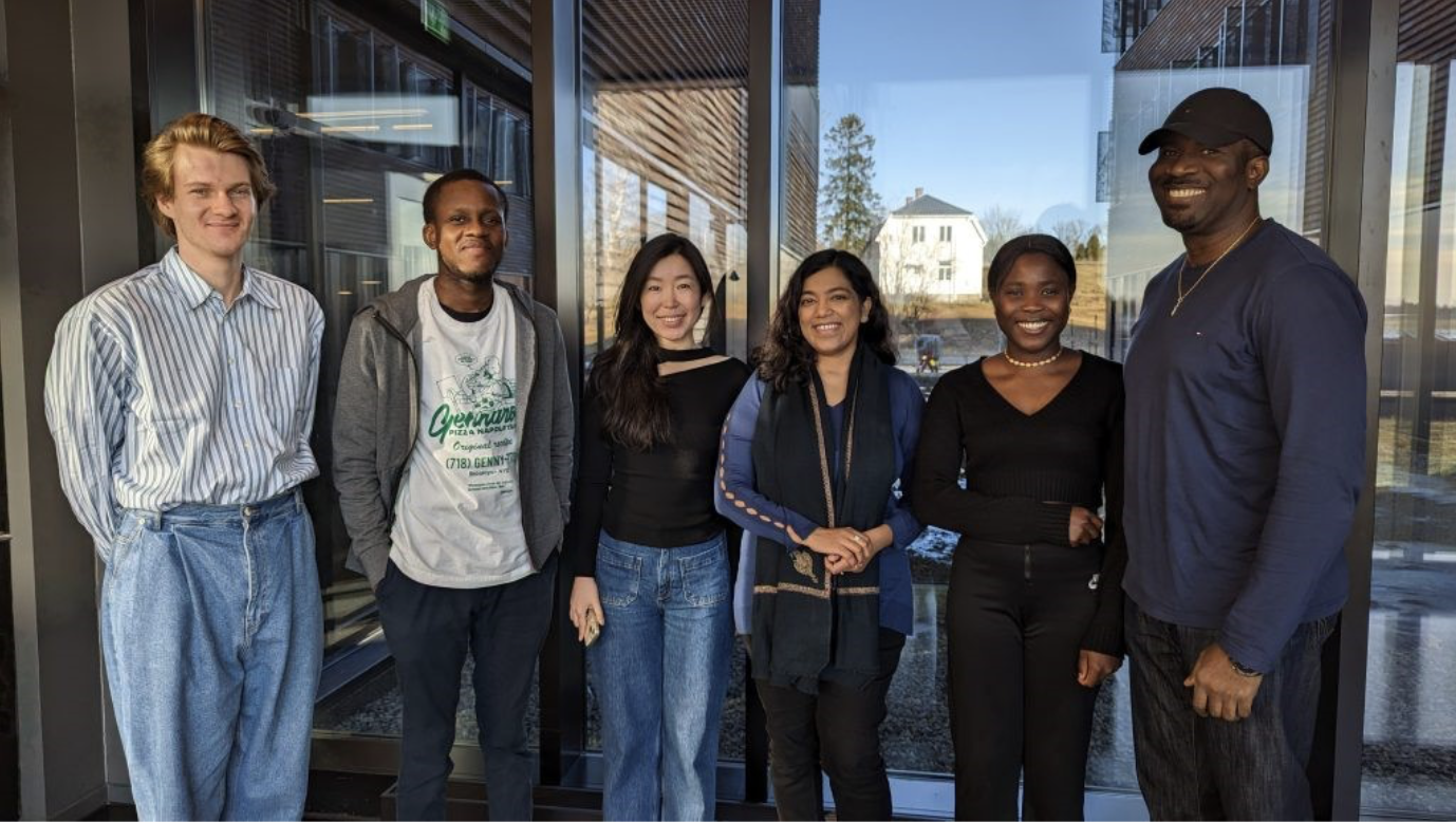 Group of six diverse people smiling inside a building with glass walls, with a view of trees and houses outside. Interns.