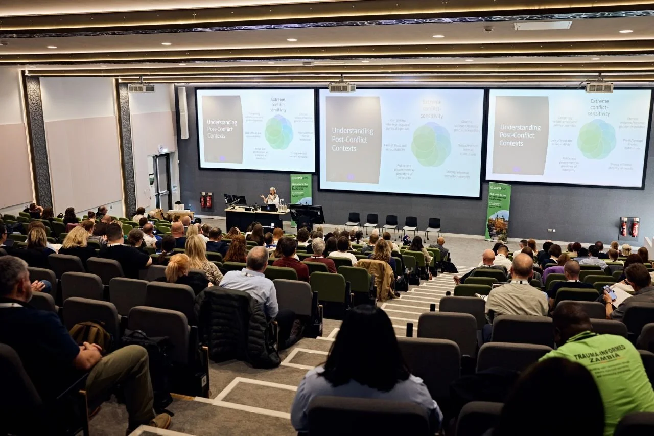 A large conference room filled with attendees facing a stage with three large screens displaying a presentation titled 'Understanding Post-Conflict Contexts.' The room has tiered seating with green and black chairs, and the lecture hall has a modern design with lighting and decorative panels on the walls.