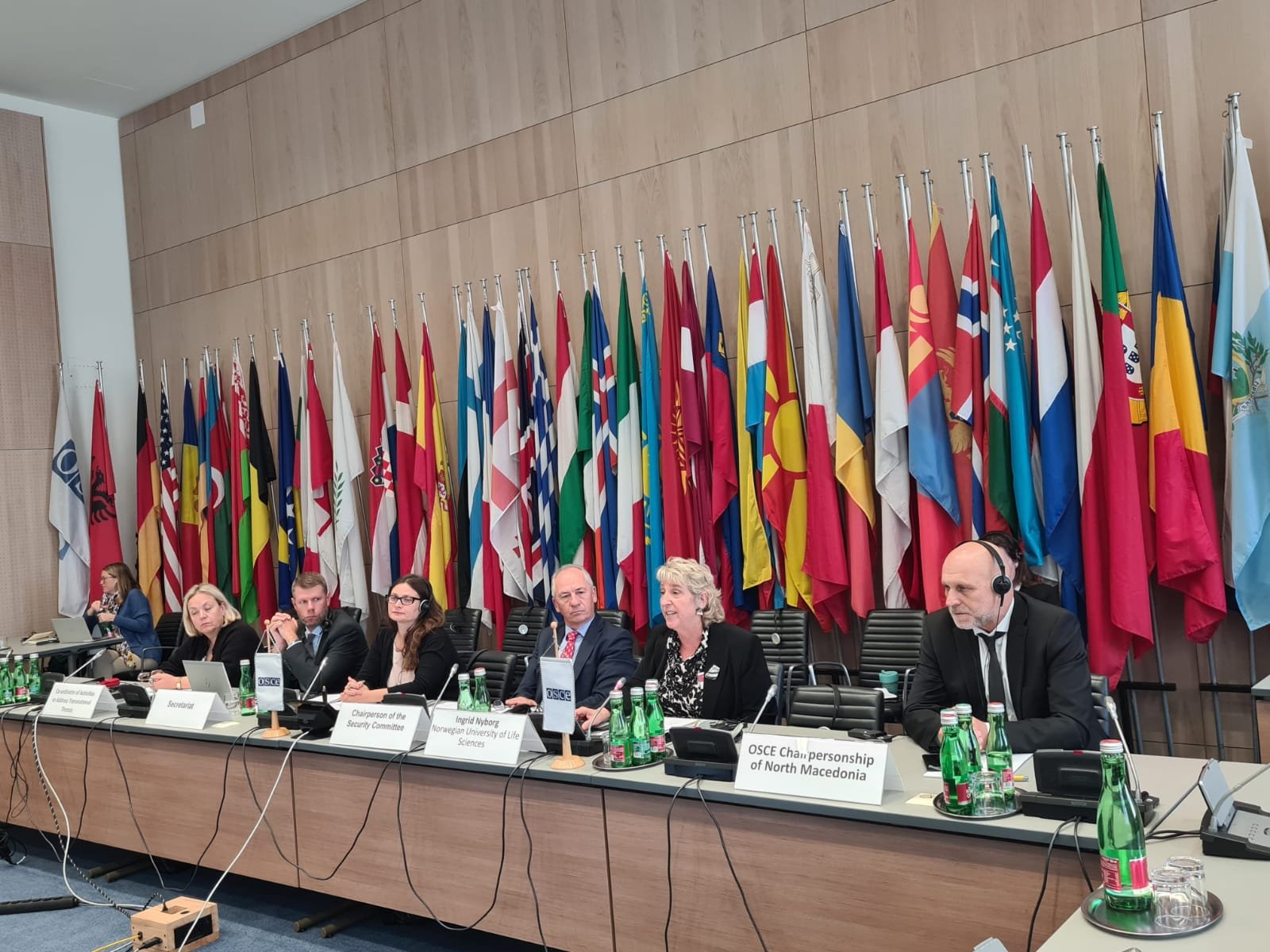 A group of five individuals seated at a long table with nameplates and bottled water, in front of a wall adorned with numerous international flags. The setting appears to be a formal international conference or meeting.