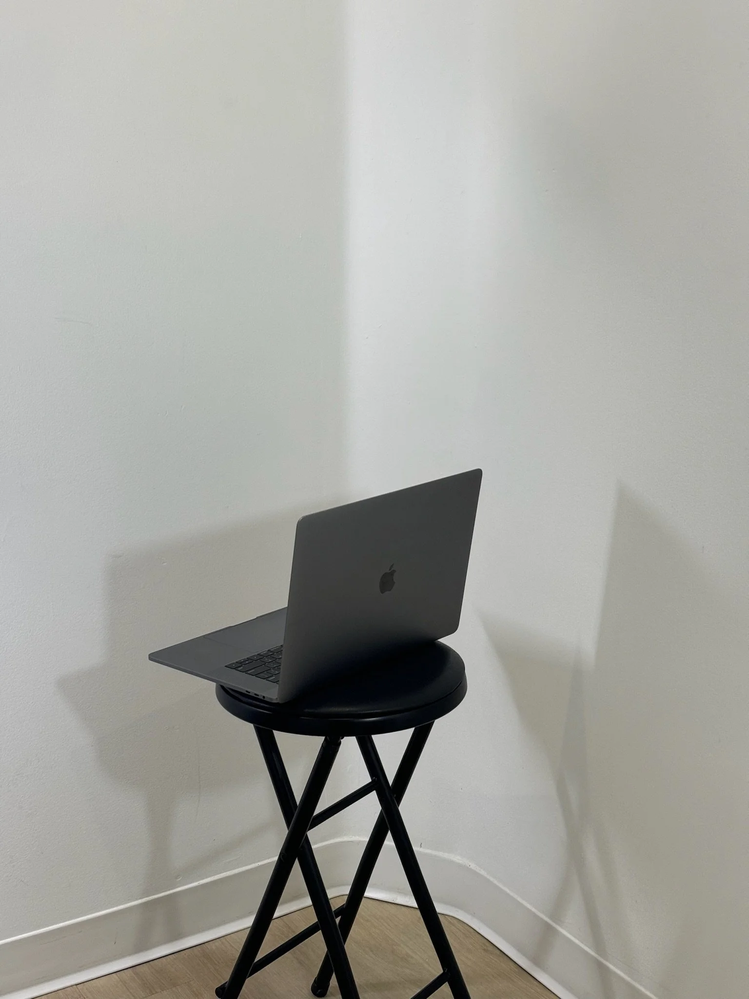 A silver MacBook laptop resting on a black round stool with crossed black legs, placed in the corner of a room with white walls and light wood flooring.