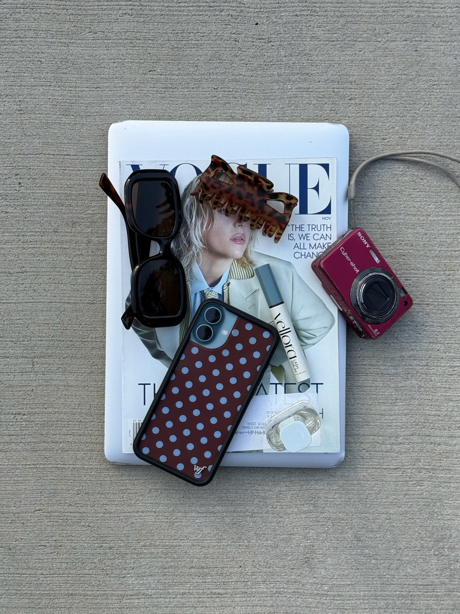 A top view of a closed Vogue magazine on a beige surface with various personal items on it, including a pair of black sunglasses, a tortoiseshell hair clip, a red compact camera, a smartphone with a red and white polka dot case, and a small bottle of perfume.