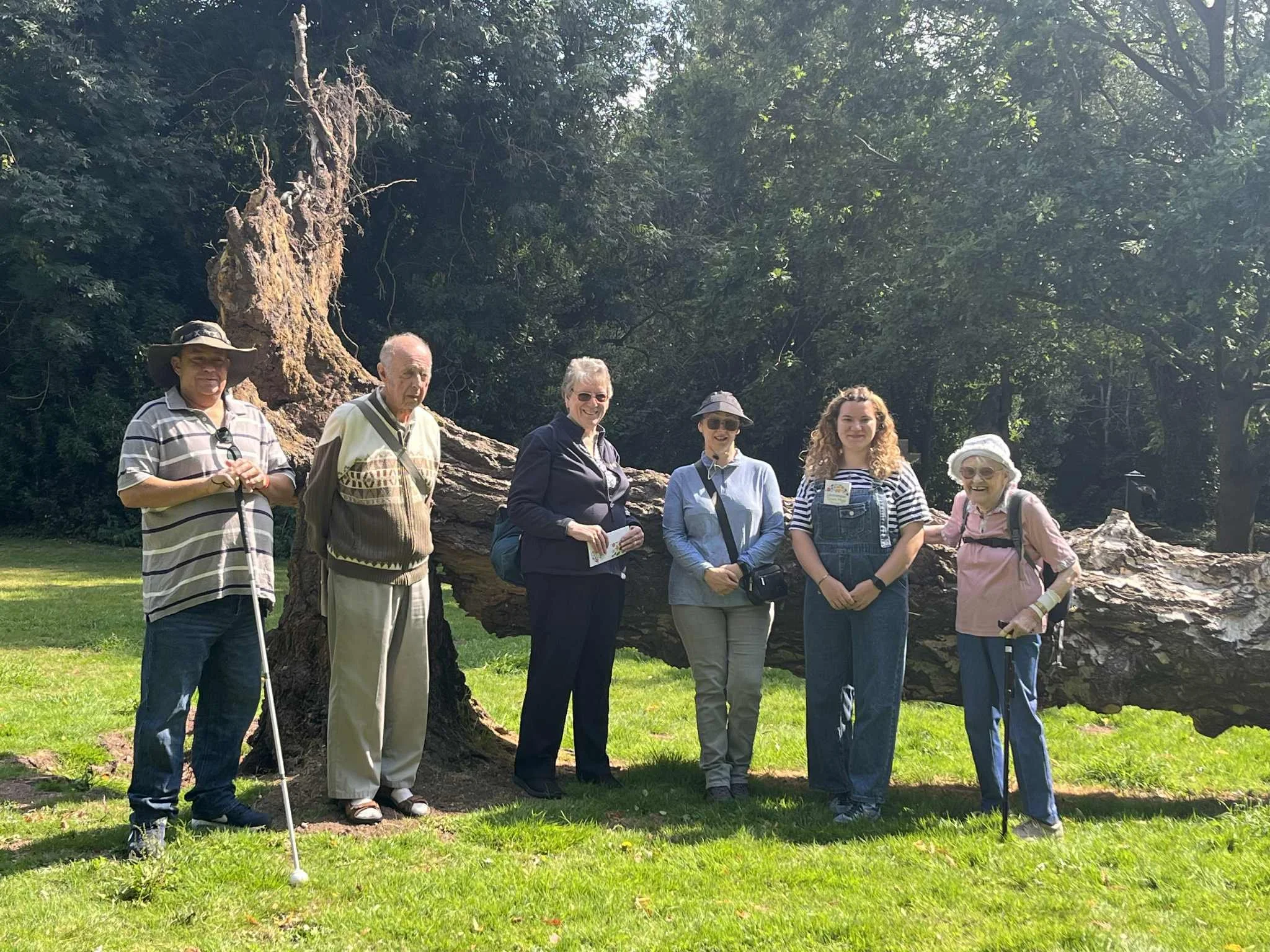 Group of six people standing on grass in front of a fallen tree, with trees and greenery in the background.