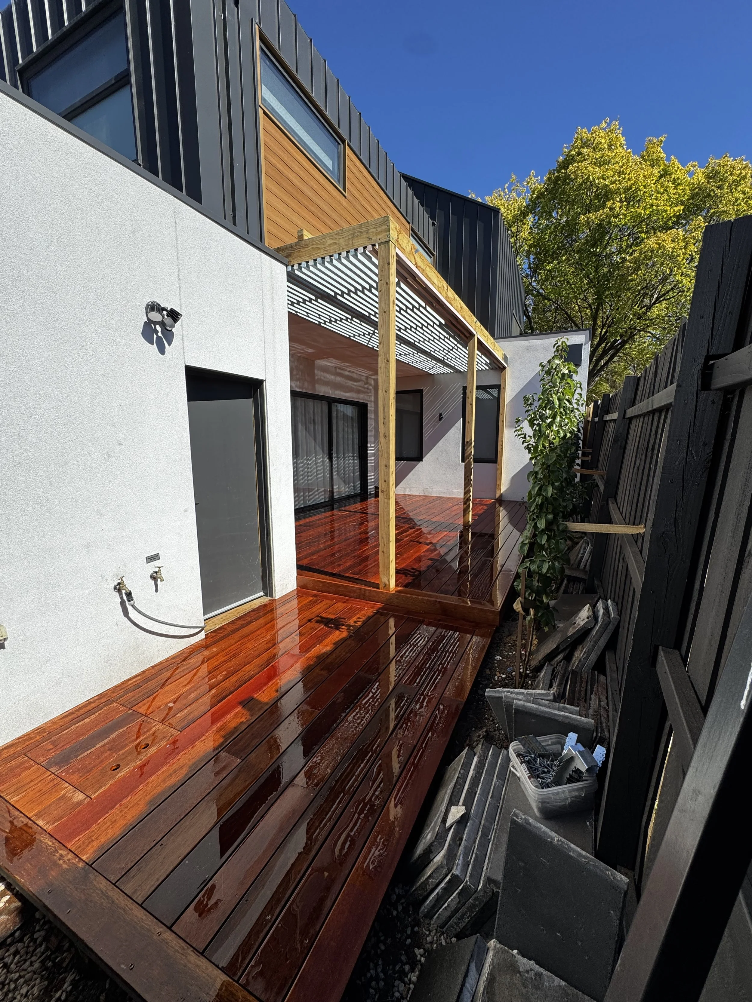A polished wooden deck surrounded by lush greenery leading to a modern house with a red door.