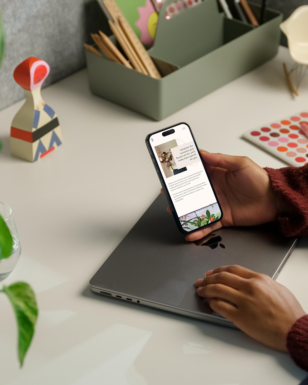 A person holding a smartphone displaying a website, with the phone resting on a closed laptop. On the desk, there's a decorative wooden figure, a container holding office supplies, a color swatch book, and leaves from a plant.