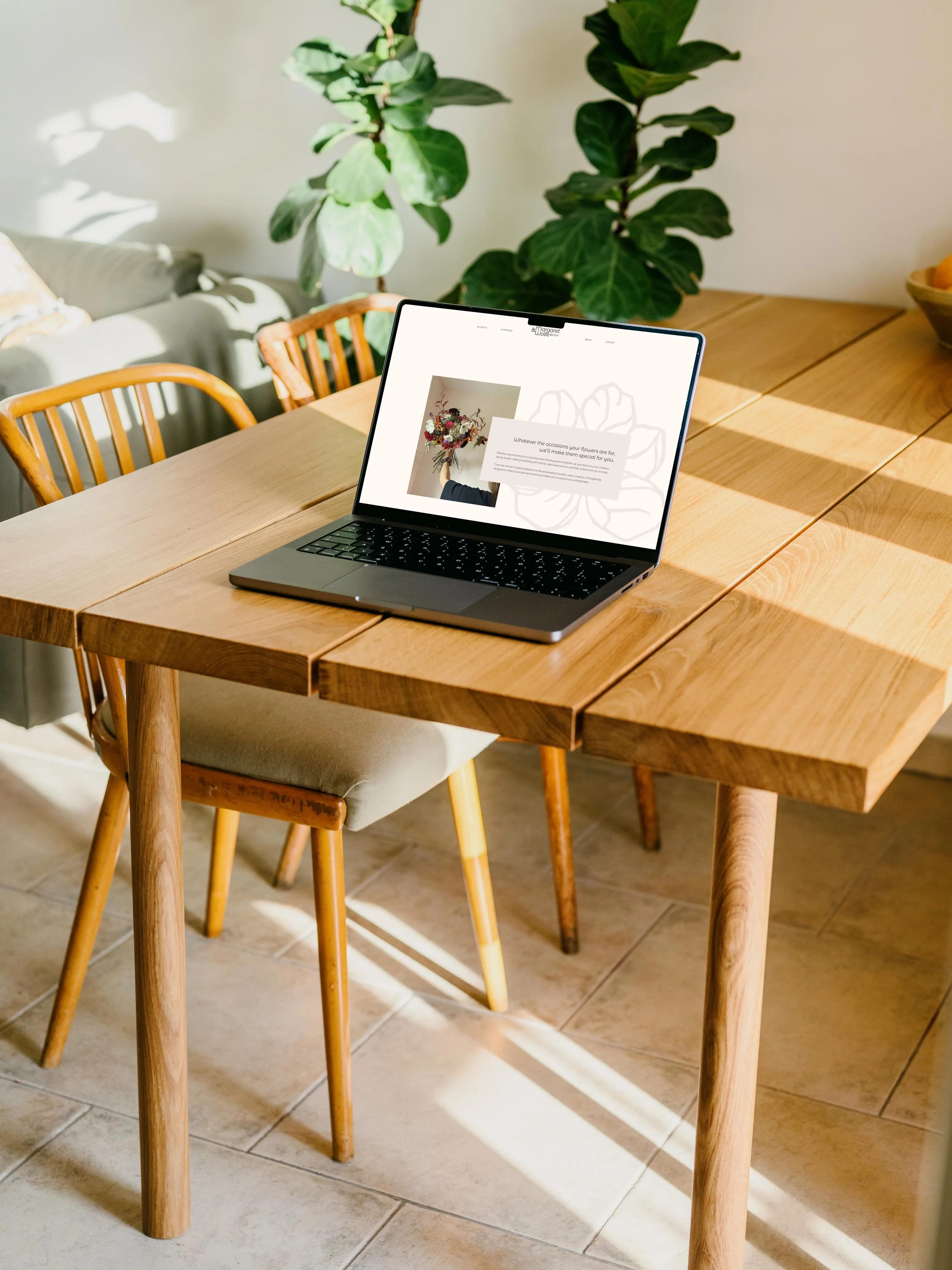 Laptop on a wooden dining table with a modern interior design, featuring plants, chairs, and natural light.