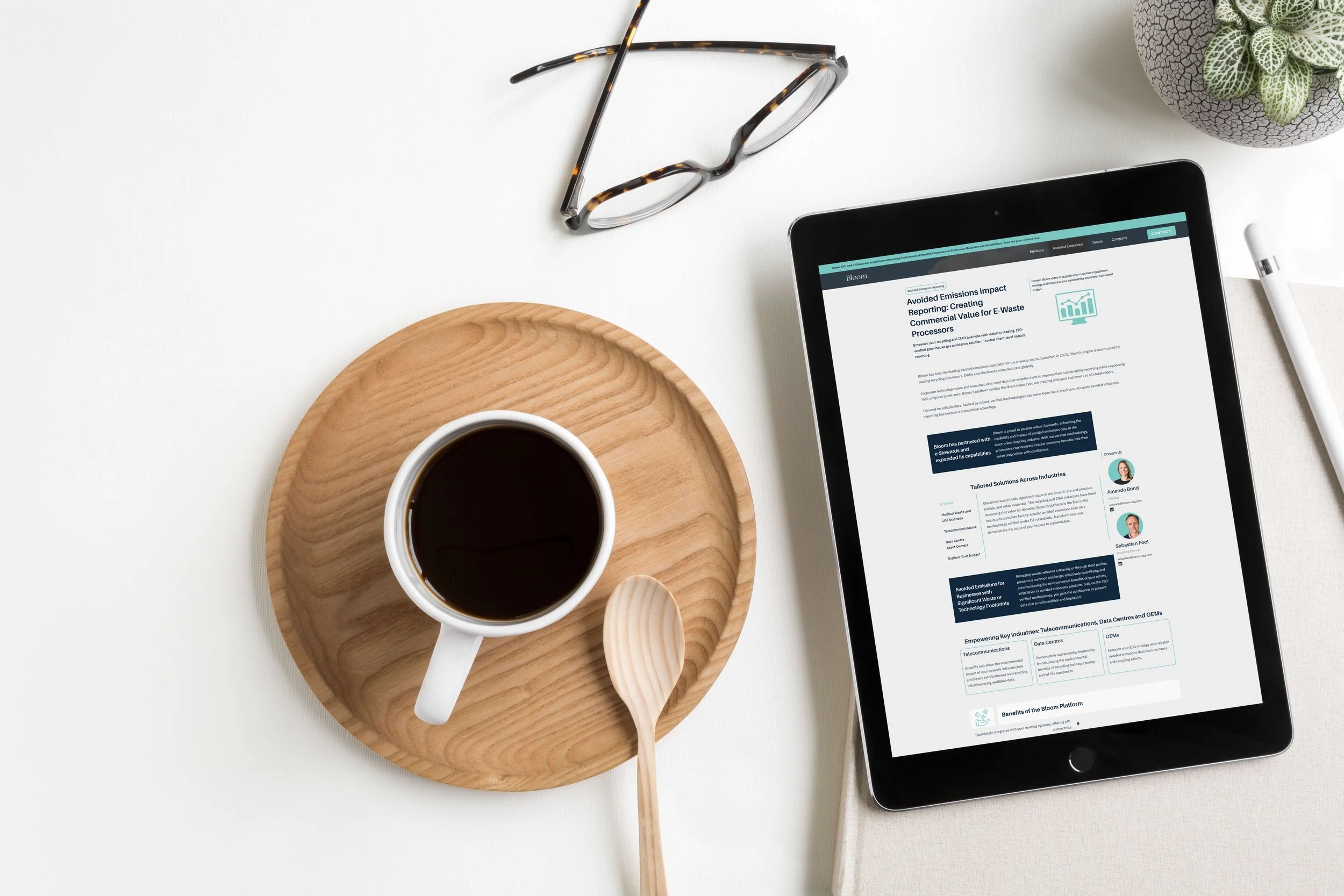 A flat lay image showing a cup of coffee on a wooden tray with a wooden spoon, next to an iPad displaying a document about emissions impact reporting. Glasses, a pen, and a small potted plant are also visible on the white table surface.