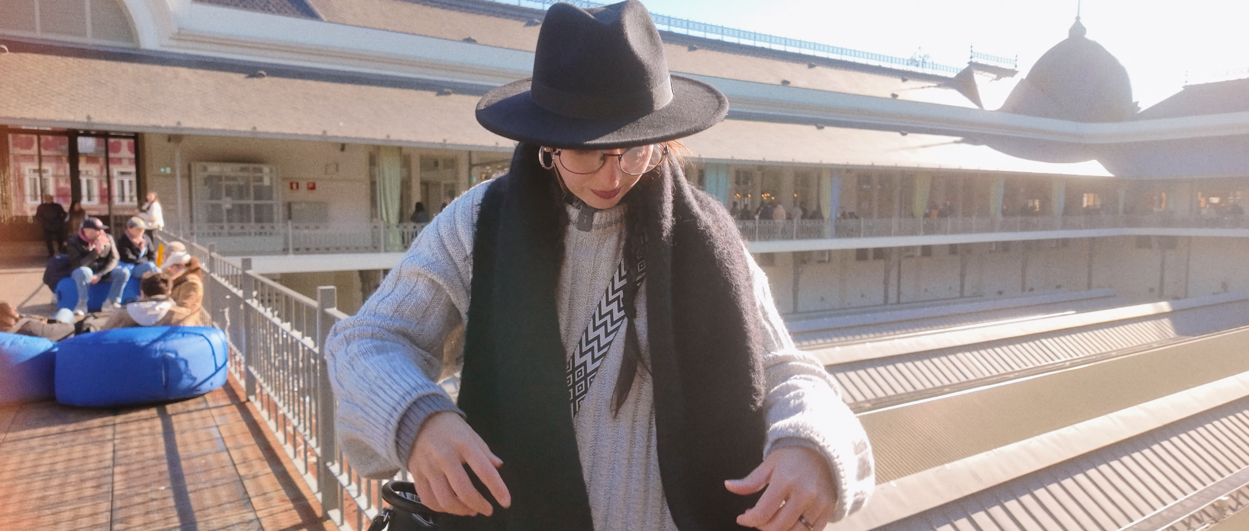 Person in black hat and glasses wearing a gray sweater and scarf, standing on a rooftop terrace with a blue bean bag and people in the background.