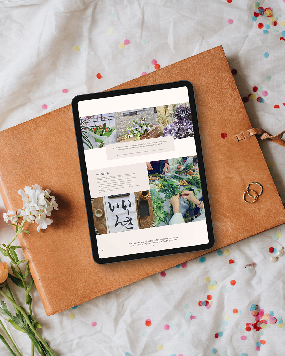 Tablet displaying a website with images of plants and greenery, lying on a tan leather book cover. White flowers and colorful confetti are scattered on a white fabric surface.