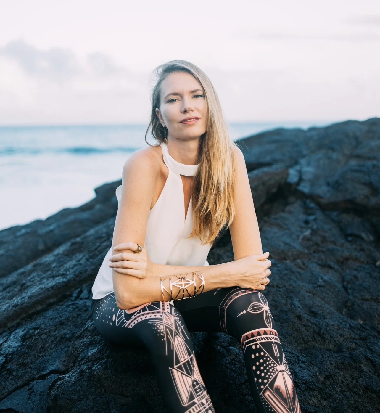 A woman with long blonde hair sitting on black rocks by the ocean, wearing a white sleeveless top and patterned leggings, with an overcast sky in the background.