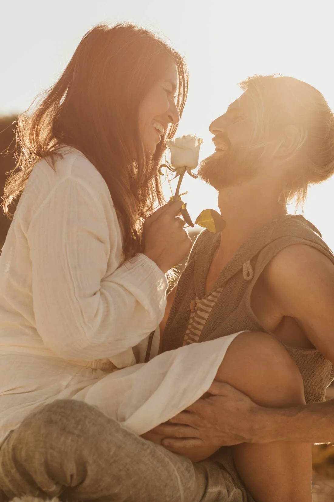 A man and woman outdoors during sunset, the woman holding a white rose near the man's nose as they smile at each other.