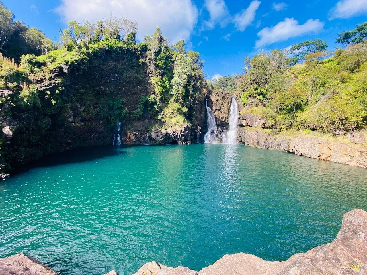 A scenic landscape featuring a turquoise lake with two waterfalls cascading from rocky cliffs surrounded by lush green trees under a partly cloudy sky.