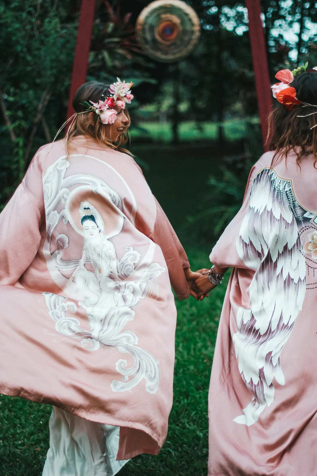 Two women wearing pink kimono robes with Asian-inspired designs, holding hands, outdoors in a green setting with a hanging wind chime in the background. Both women have floral headpieces.